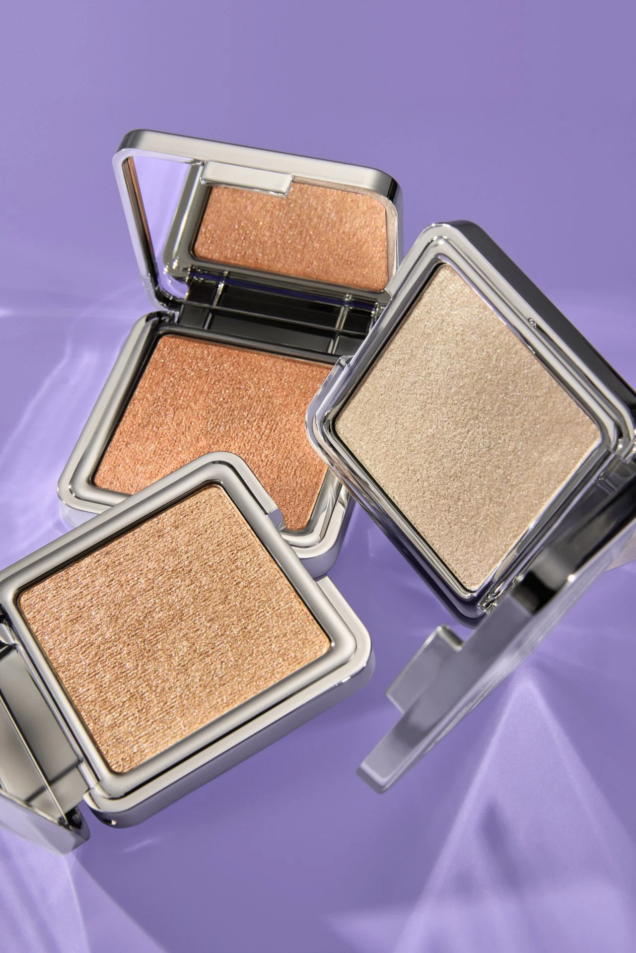 Multiple highlighter makeup compacts with shimmery shades in silver cases on a purple background.