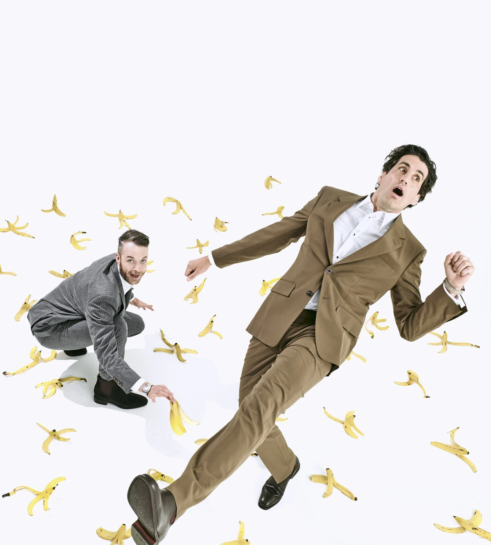 Two men in suits falling or kneeling among scattered banana peels on a white background, with surprised or shocked expressions.
