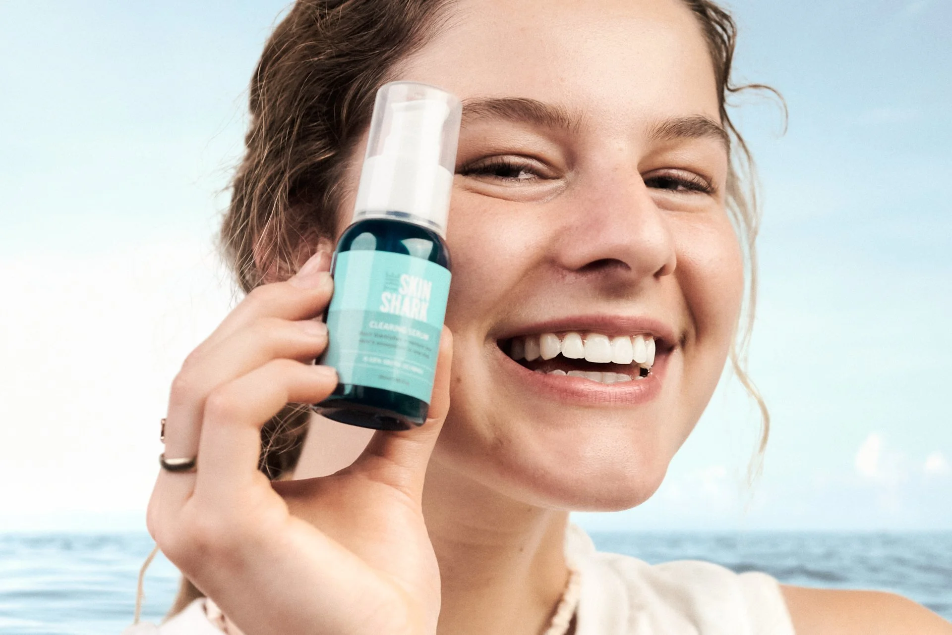 A happy young woman smiling and holding a bottle of skincare product labeled 'Skin Shark' near her face against a beach background.