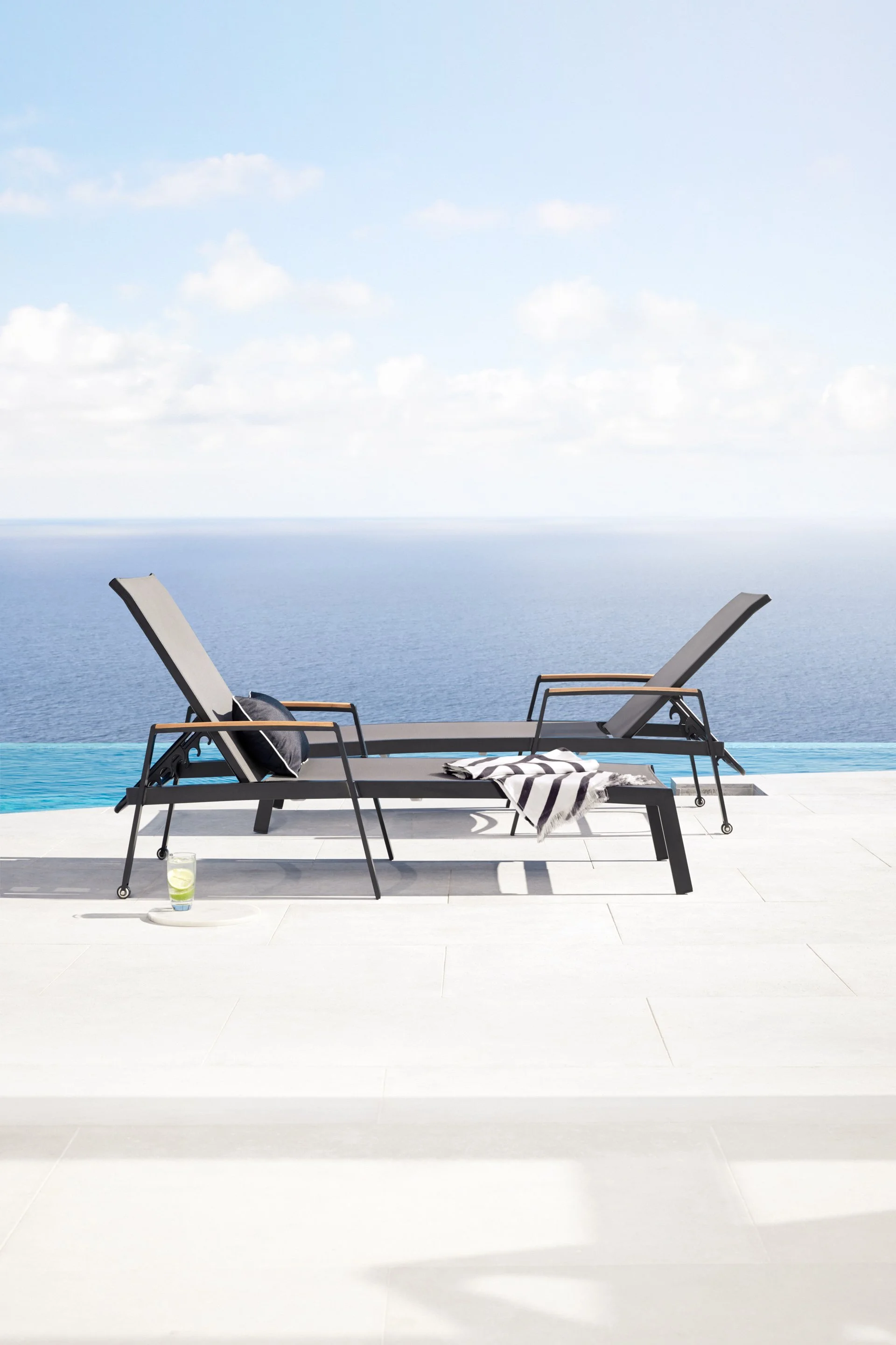 A luxurious infinity pool overlooking the ocean with two black lounge chairs, a striped towel, a glass of lemon infused water, and an umbrella cast shadows on the white tiled pool deck against a cloudy sky.