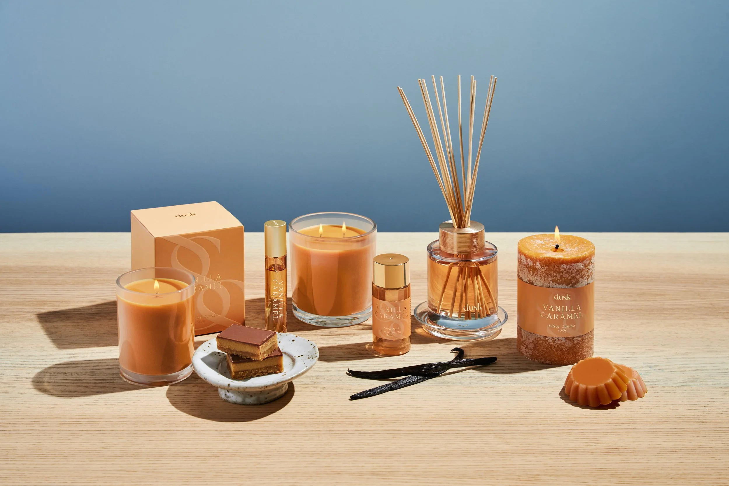 A collection of vanilla caramel scented candles, reed diffuser, baked treats, vanilla and caramel scented products, and a slice of cake on a wooden table against a blue wall.