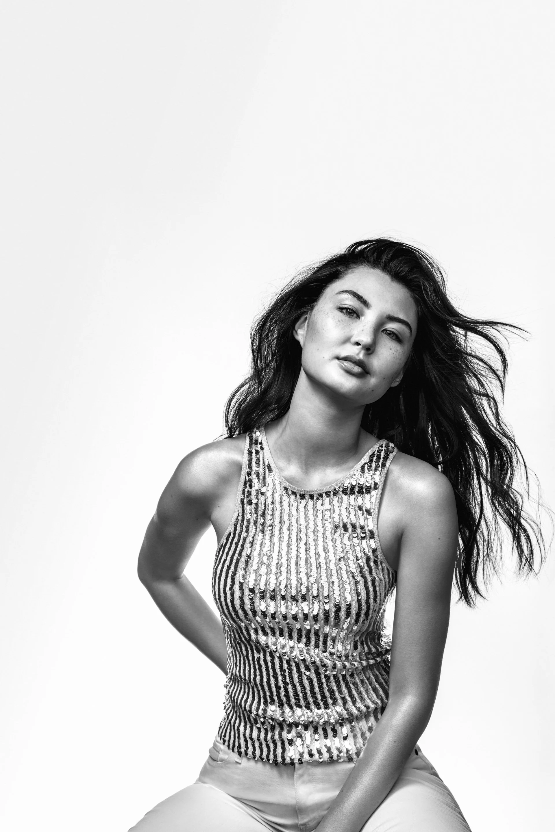 A black and white photo of a woman with long, wavy hair looking at the camera. She is wearing a sleeveless, sequined top and light-colored pants, with a confident expression and a slight head tilt.