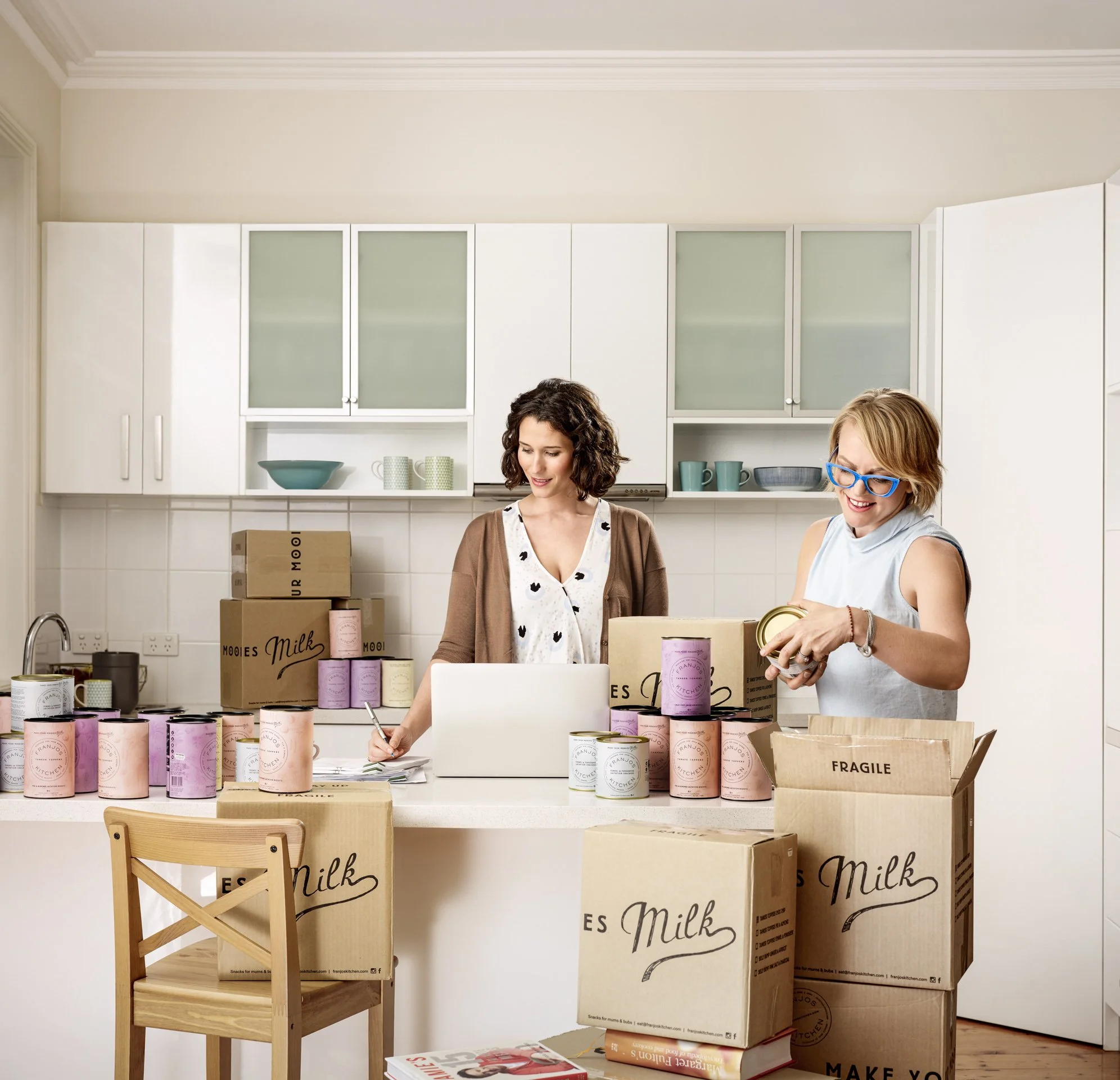 Two women packing food items in a kitchen, surrounded by boxes labeled 'Milk' and 'Fragile'.