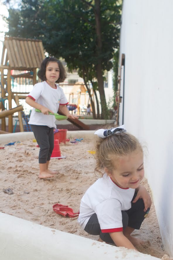 Duas alunas brincam em um tanque de areia na área externa da escola, atividade inspirada na abordagem da Criança Reggio, em prática na escola.
