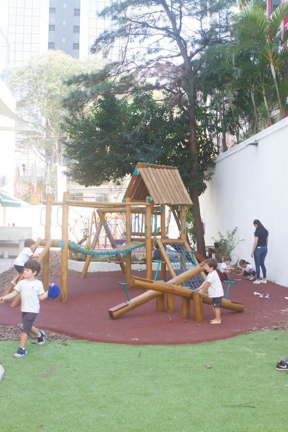 Alunos brincam no playground externo da escola que possui pavimentação segura e área de gramado natural.