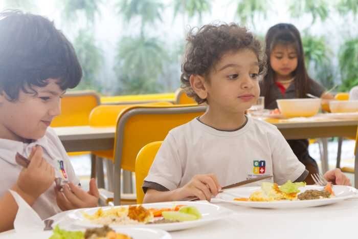 Alunos almoçando no refeitório da escola. O espaço tem mesas e cadeiras e recebe iluminação natural.