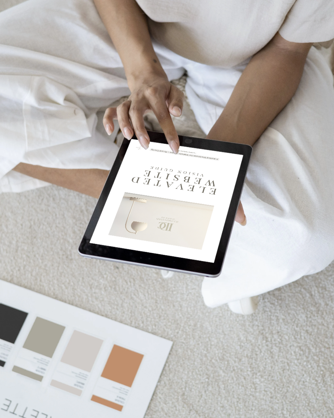 Person holding a tablet displaying an interior design book with decorative items, sitting on a white couch with a color palette chart nearby.