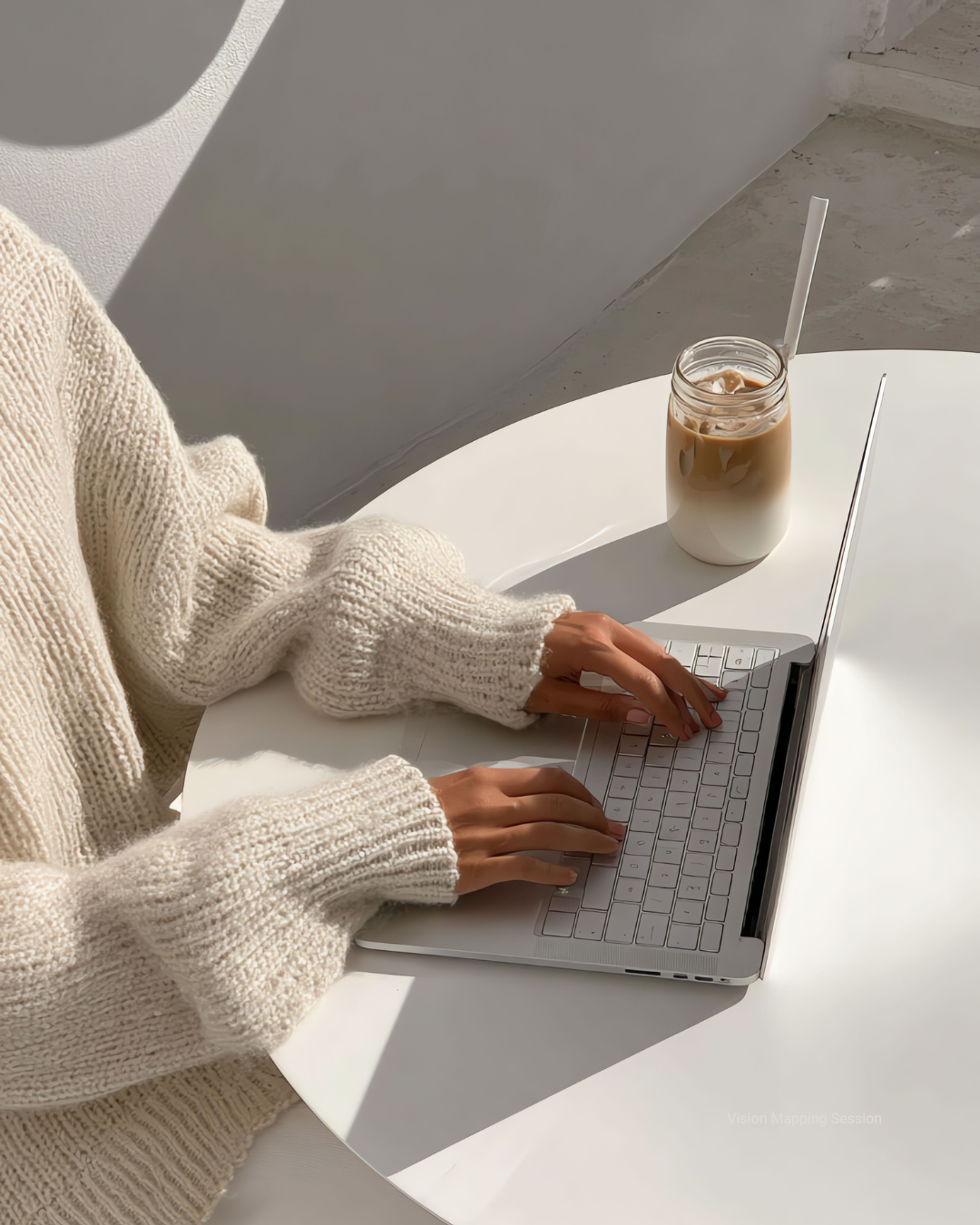 Person wearing a cream knitted sweater typing on a silver laptop on a white table. Next to the laptop is a jar of iced coffee with a straw.