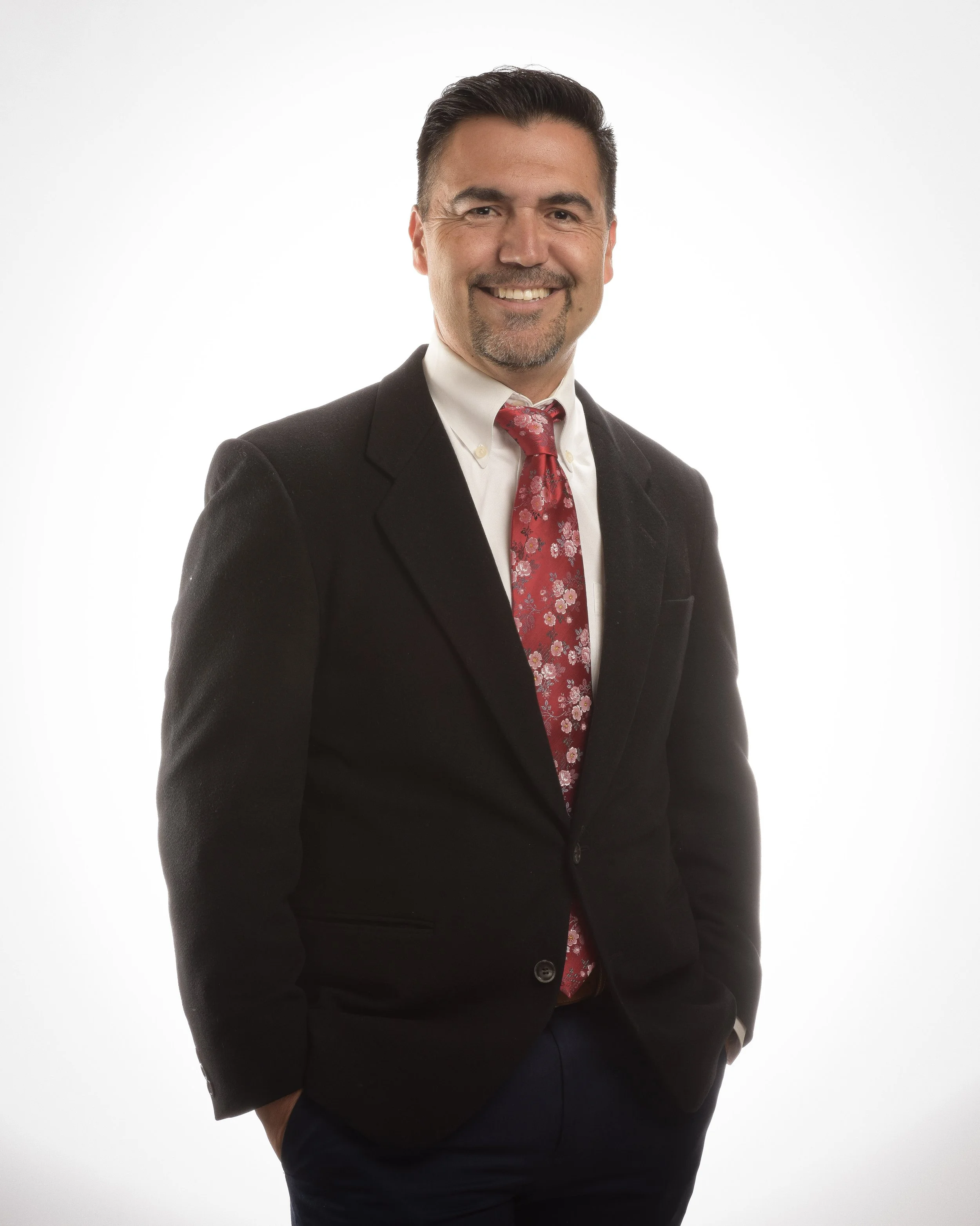 A middle aged man with black hair. He is wearing a suit and tie.