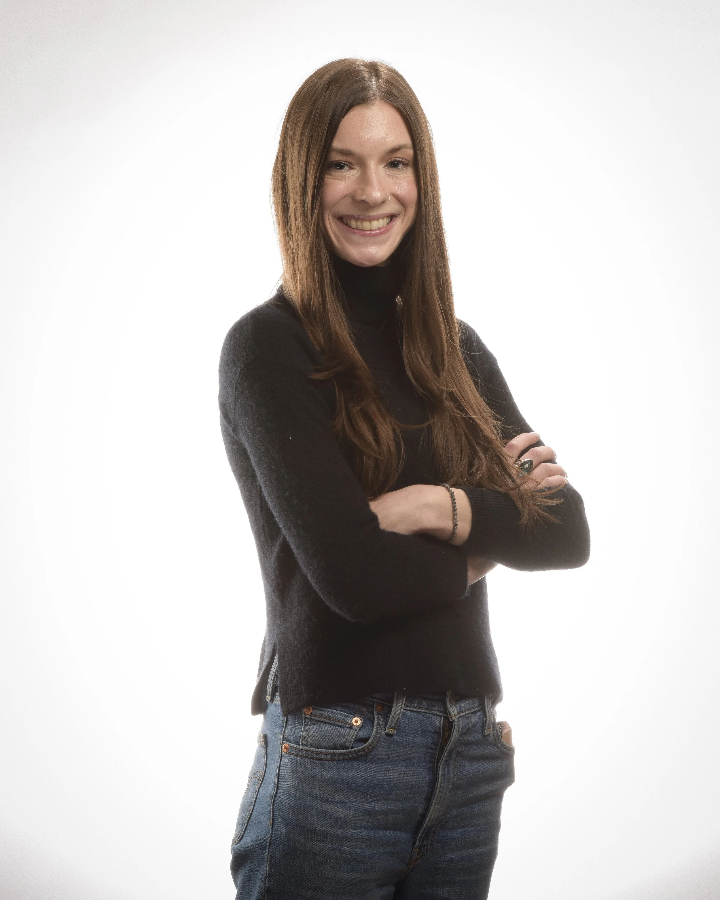 A woman with long brown hair. She has her arms crossed across her chest and is wearing a black turtle neck sweater.