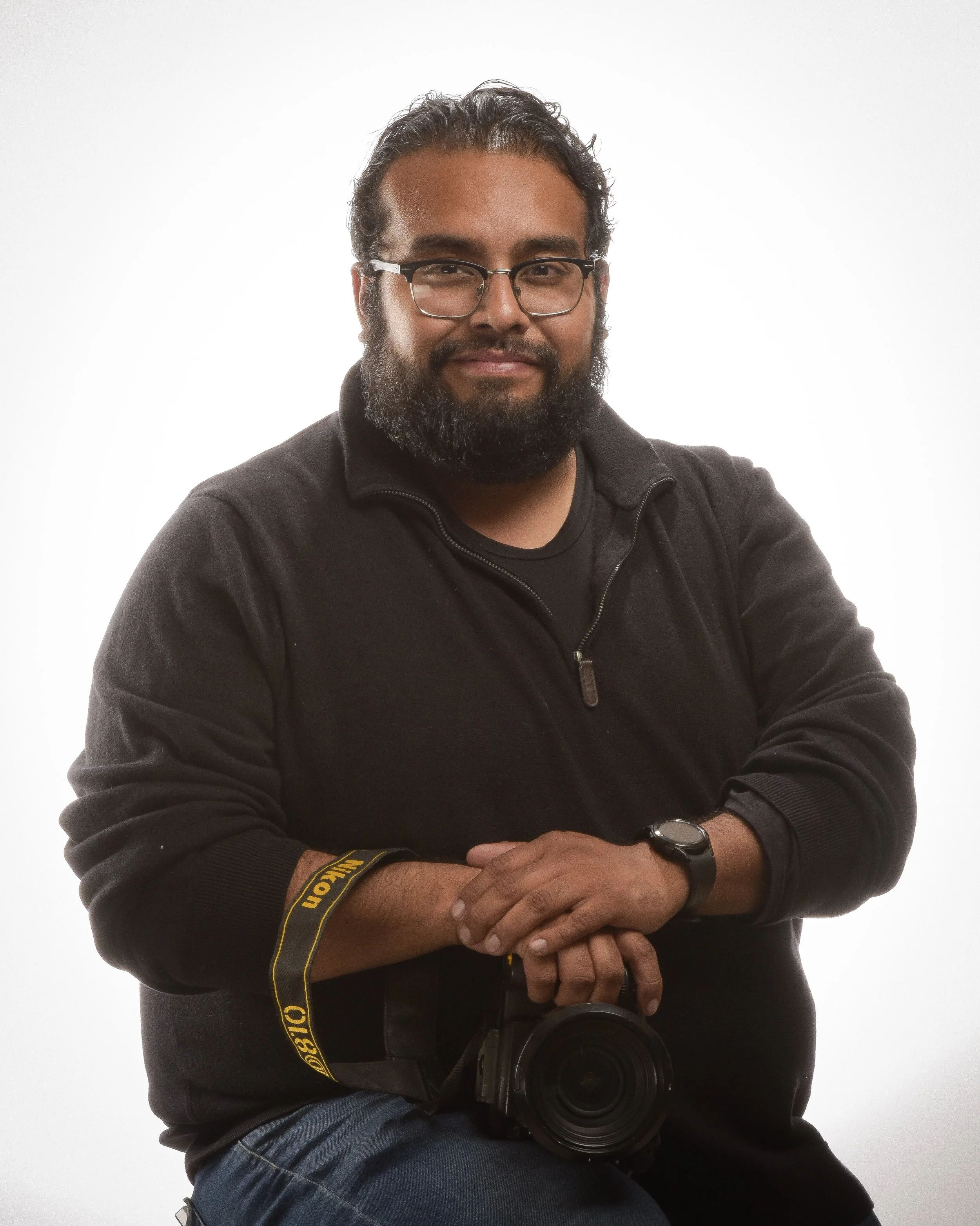 A young man with curly black hair and a thick beard. He is wearing a black sweater.