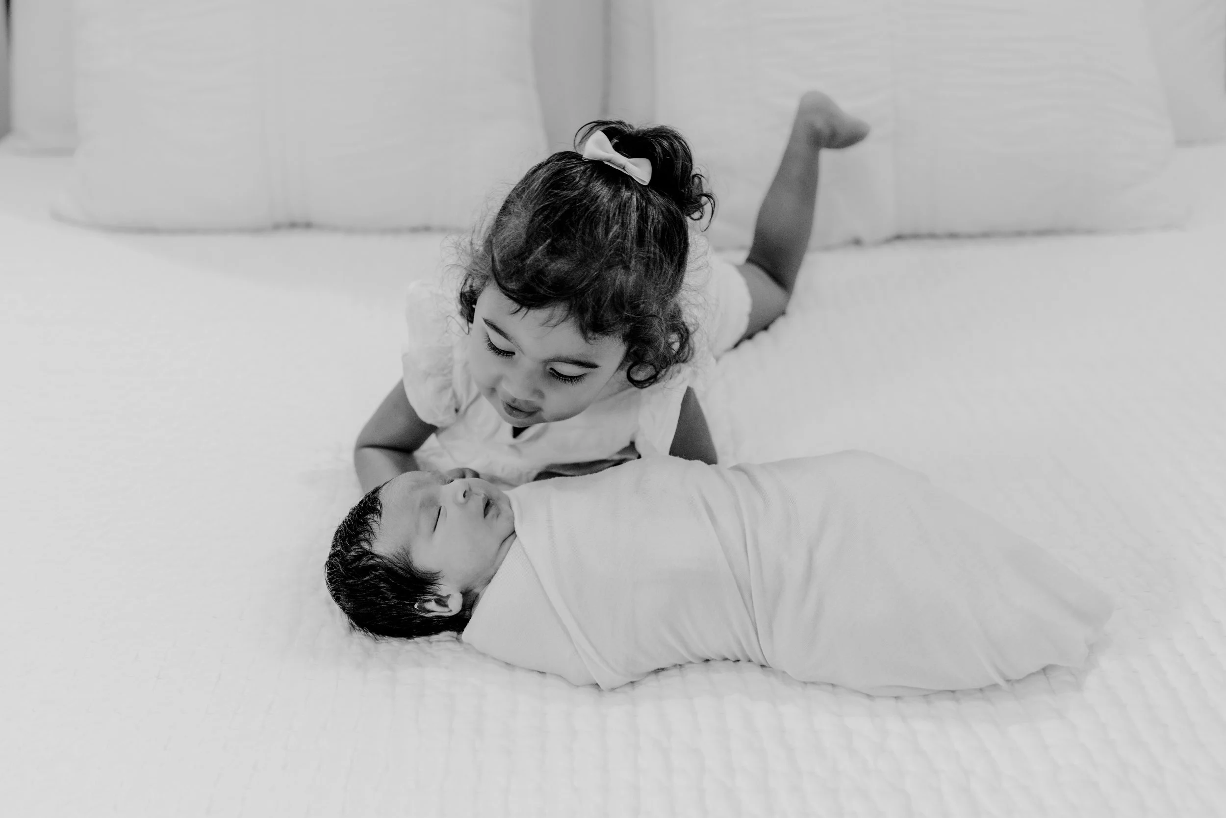 big sister curiously leaning over to look at newborn baby brother on a bed