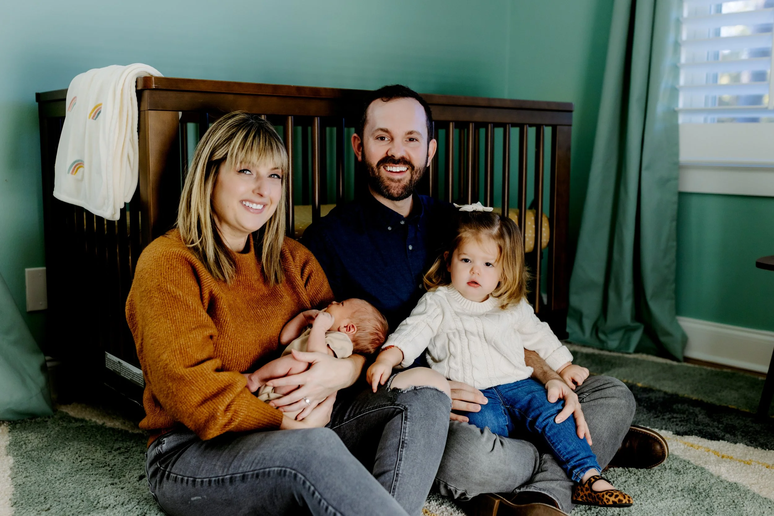 Family cuddling on floor in front of baby’s crib during natural light newborn photos at home