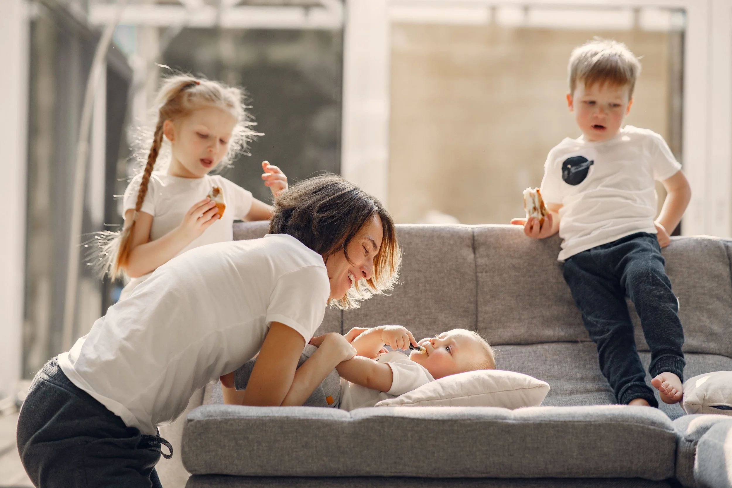 Mother smiling at her youngest child on a sofa while two older children enjoy snacks nearby.