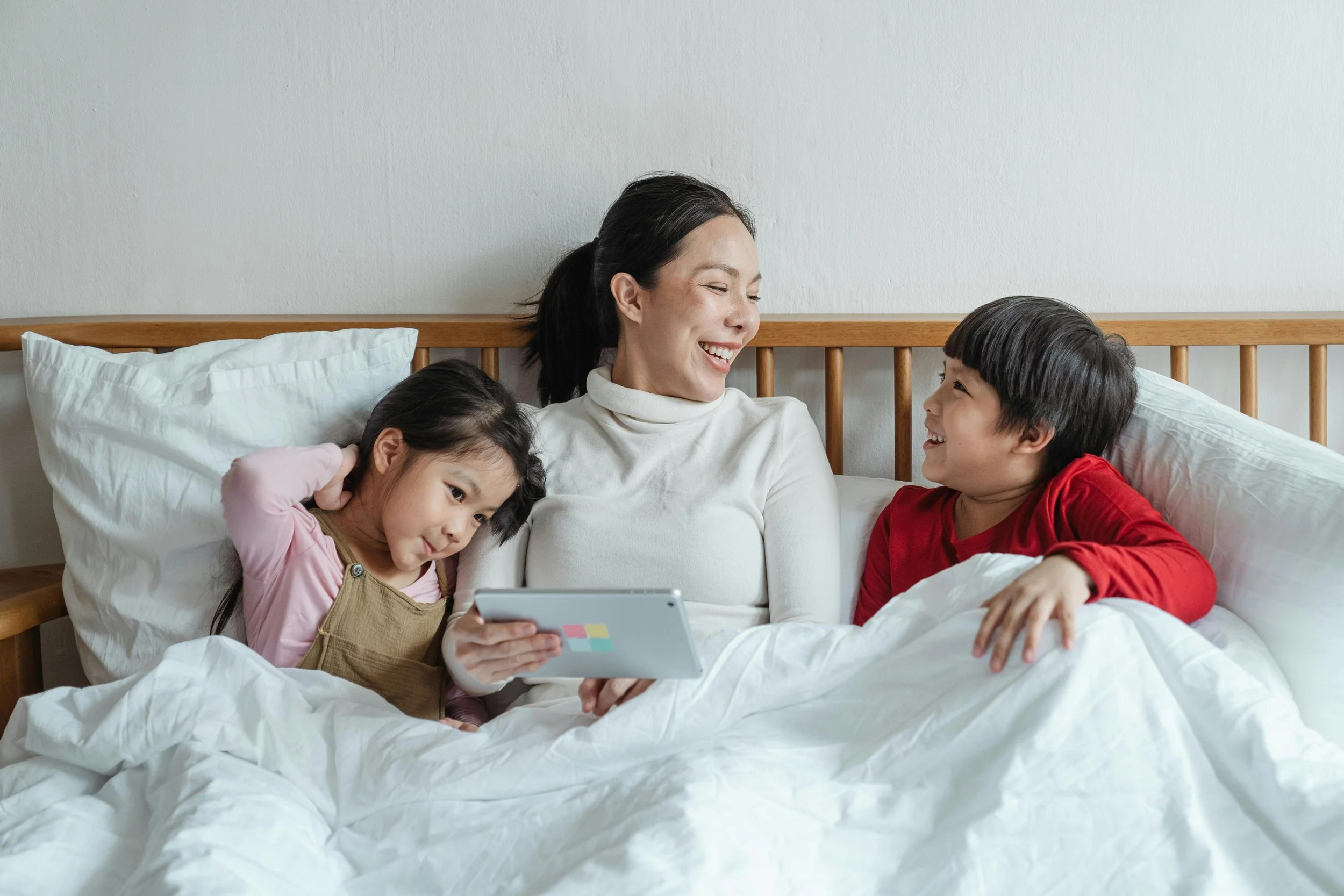 Mother and two children smiling together in a tidy bed, looking at a tablet in a calm, clutter-free bedroom.