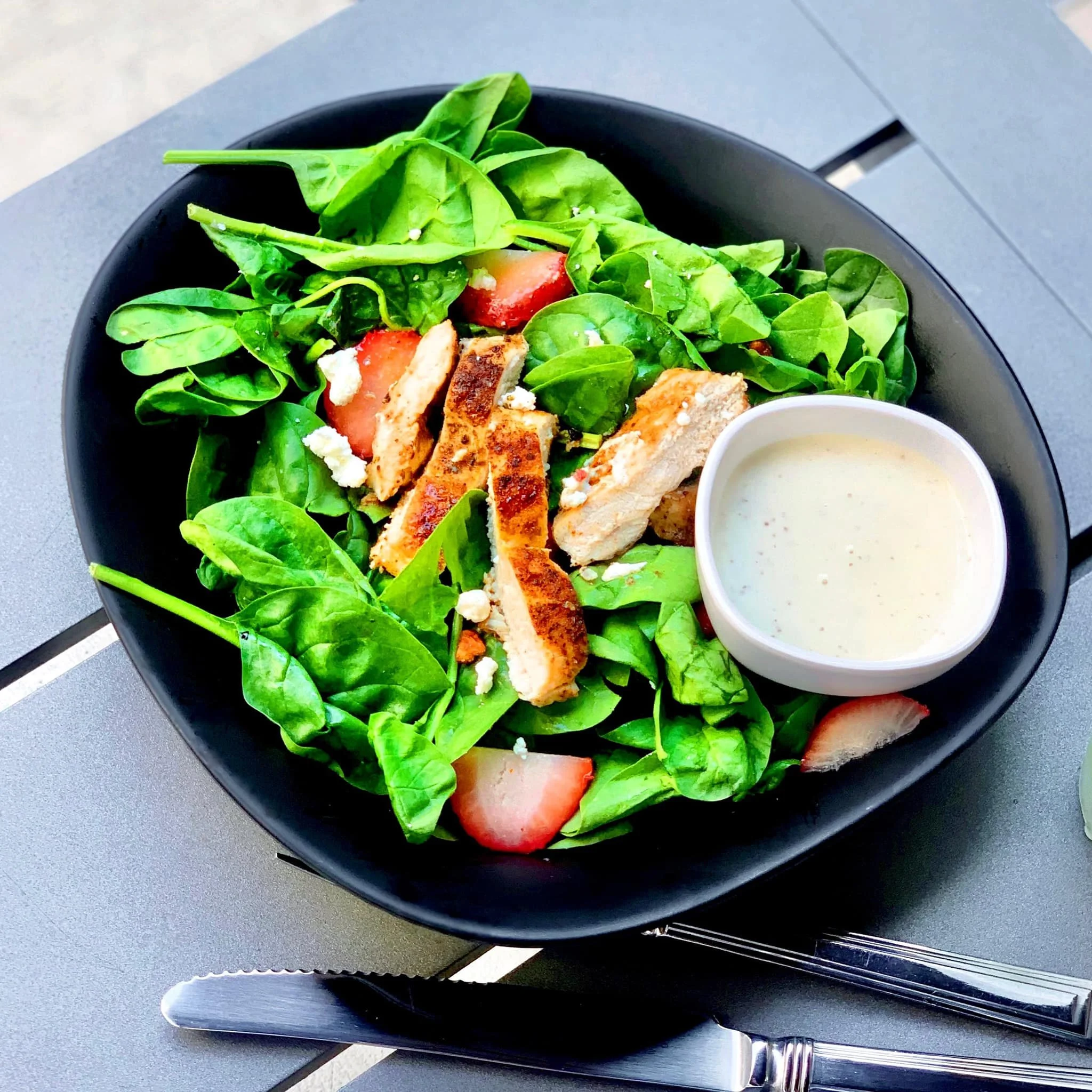 Fresh spinach salad with grilled chicken strips, sliced strawberries, feta cheese, and a side of creamy dressing in a bowl, served on a gray surface with silverware.