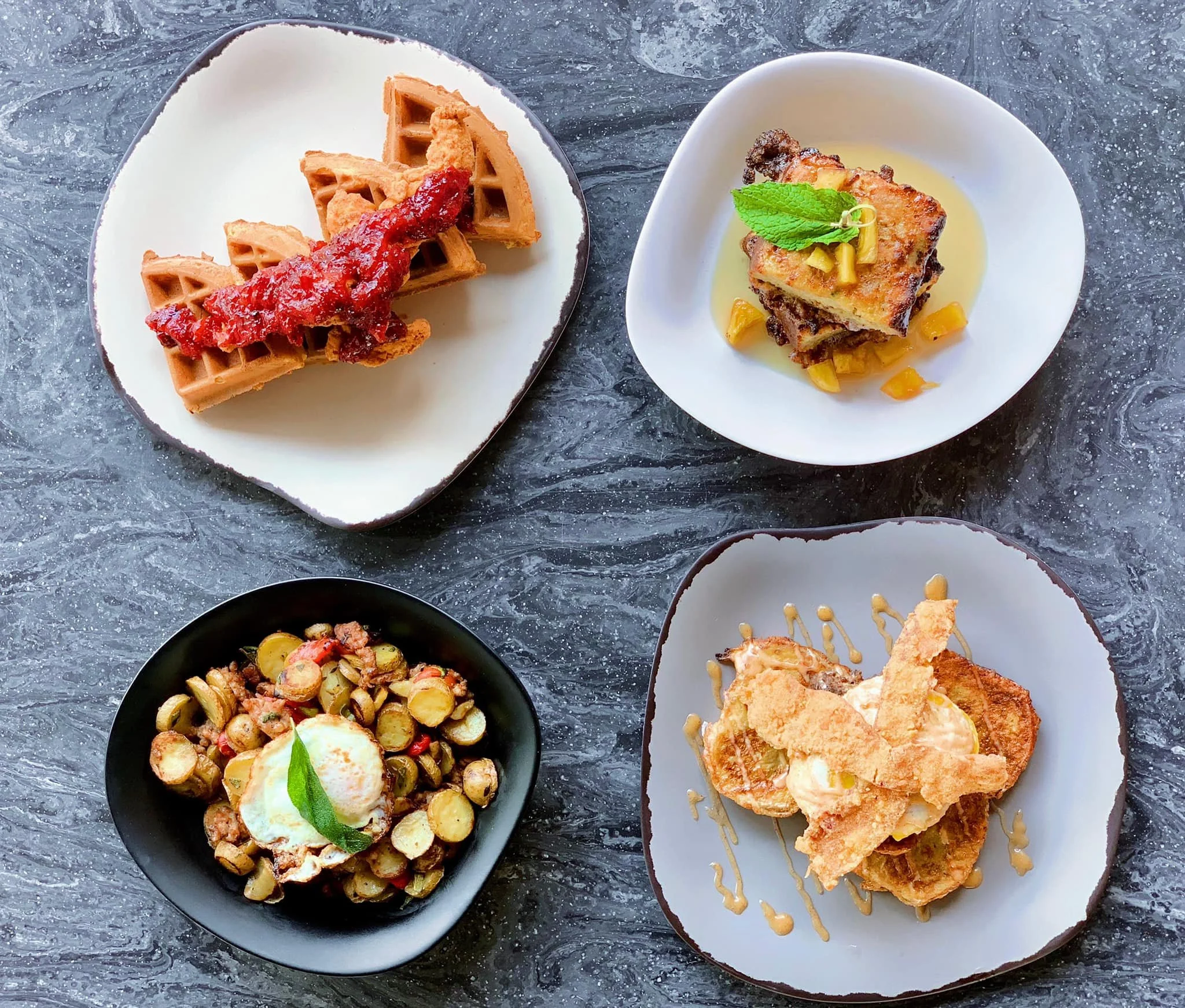 Four plates of different gourmet dishes on a dark marbled surface, including waffles with syrup, a layered dish with sauce and garnish, a bean and vegetable salad with a poached egg, and a fried fish fillet with sauce and garnishes.