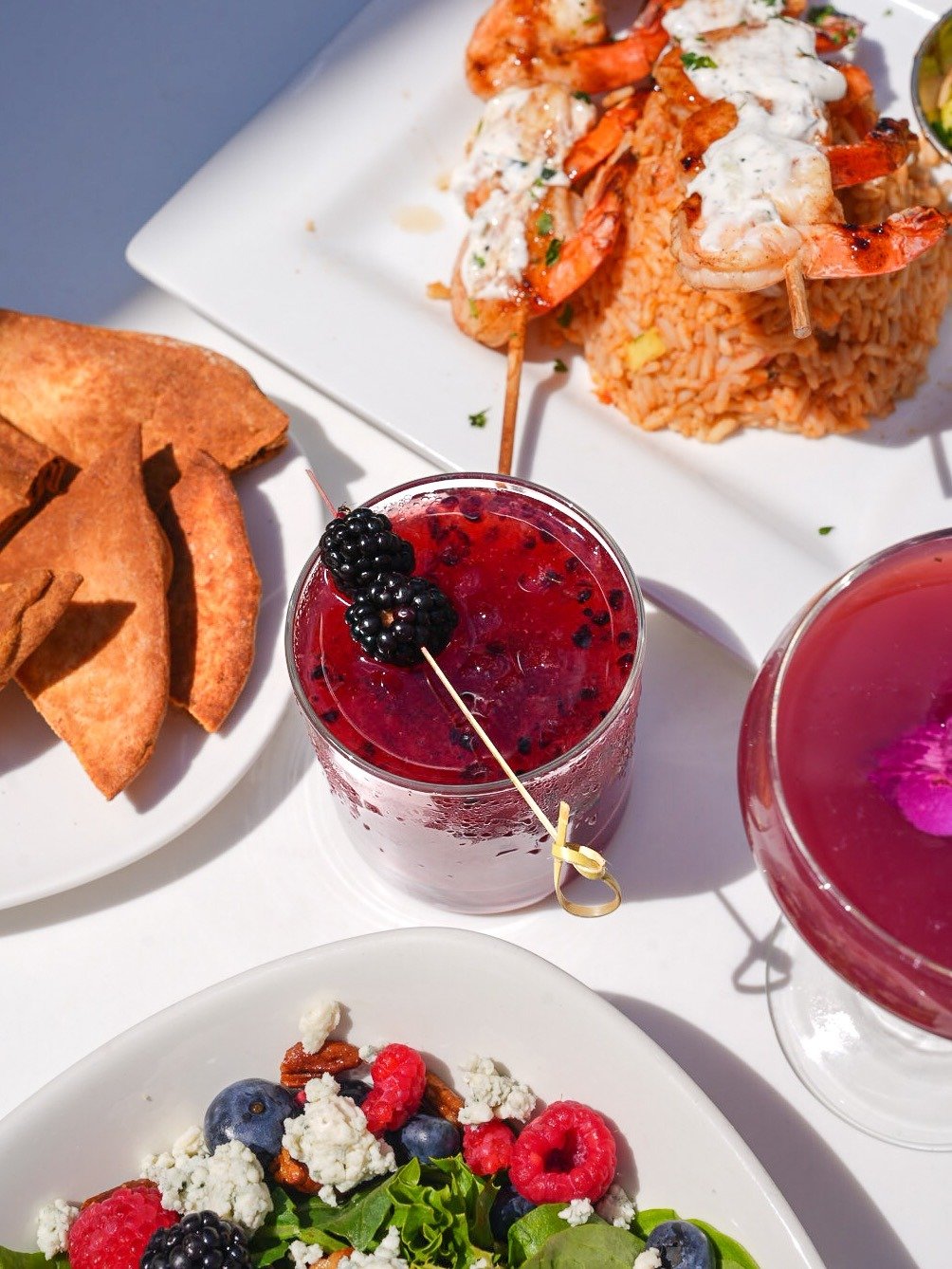 A plate of fried plantains, a glass of red berry cocktail garnished with blackberries, a plate of shrimp with rice, and a glass of pink-colored cocktail with an orchid flower, on a white table.