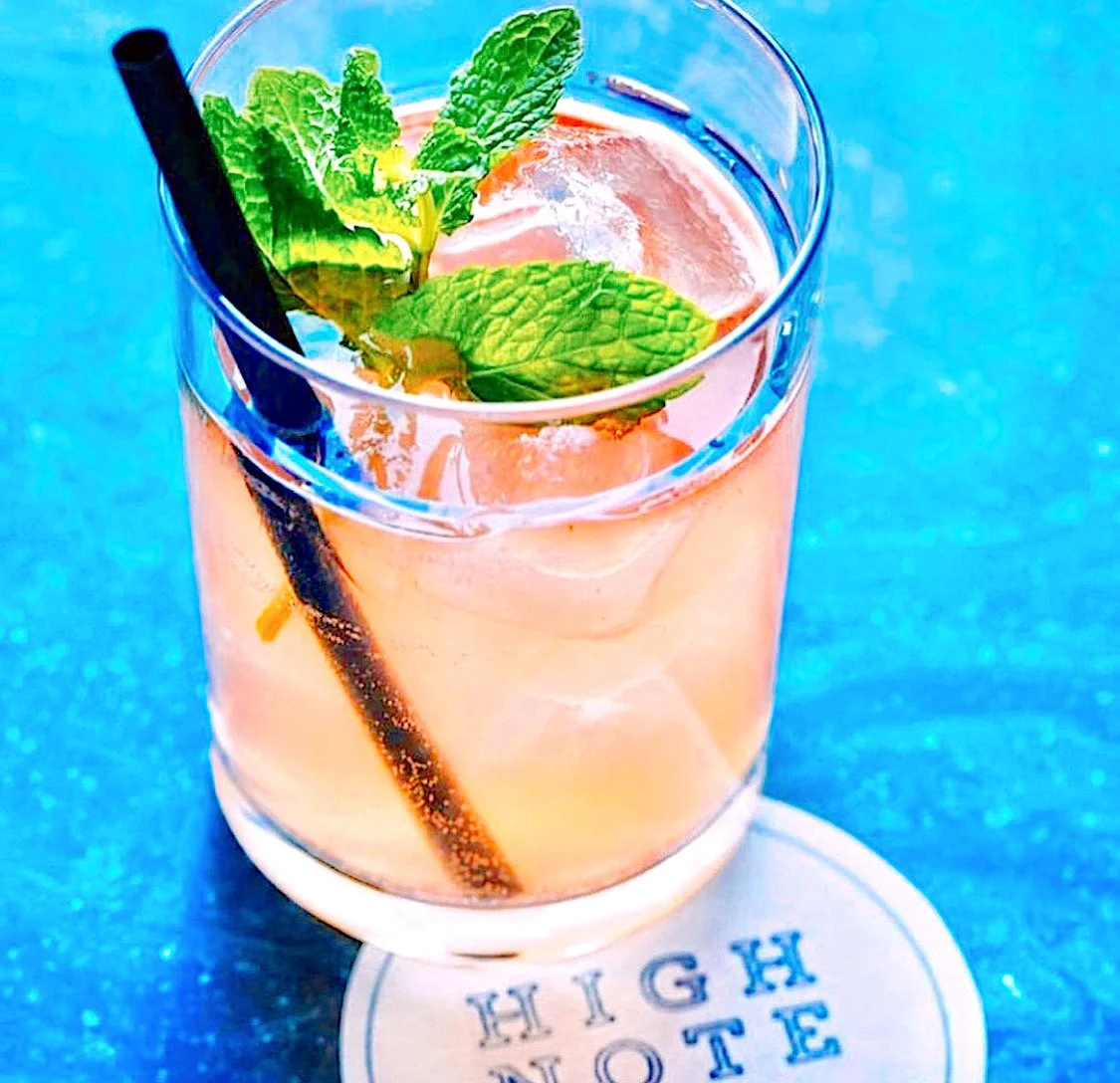 A glass of pink cocktail garnished with fresh mint leaves and a black straw, sitting on a coaster with the words 'HIGH NOTE' printed on it.