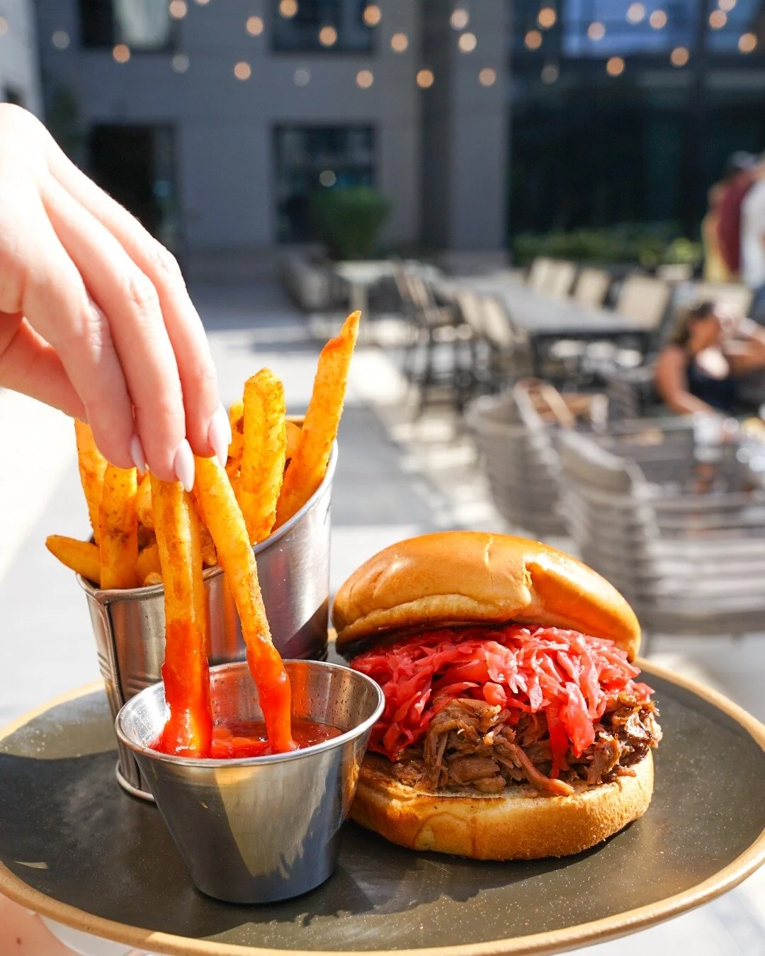 A plate with French fries, a small cup of ketchup, and a sandwich with shredded meat and pickled red onions in a bun, on an outdoor table with blurred chairs and people in the background.