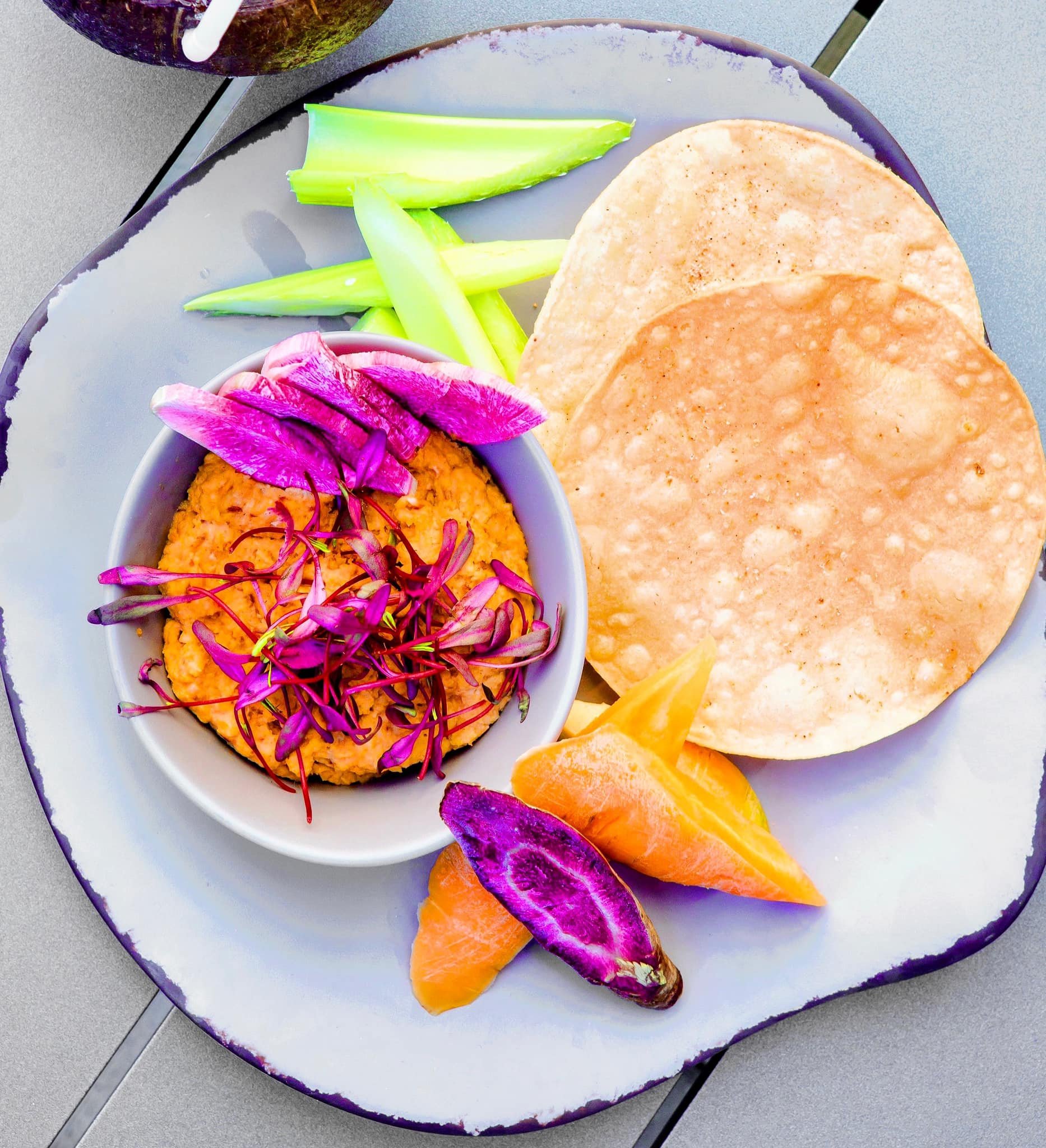 A plate with two rice cakes, a small bowl of hummus topped with purple microgreens and pink edible flower petals, and fresh vegetable slices including celery, mandarin orange segments, and purple yam slices.
