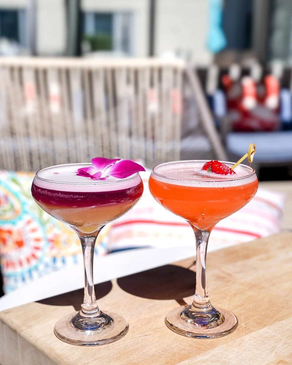 Two cocktails on a wooden surface, one with a pink flower and the other with a strawberry garnish, in an outdoor setting.
