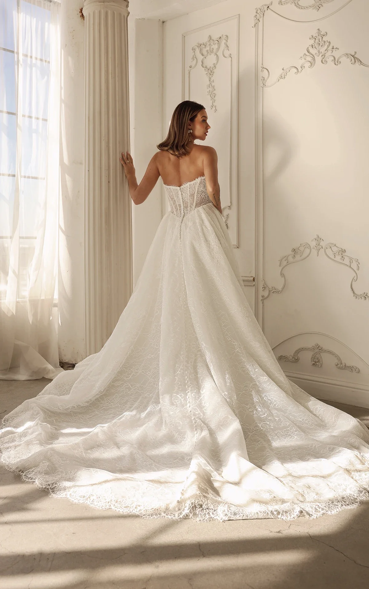 Bride/Model is provincial style room viewed from the back in a strapless lace down with a basque waist and exposed boning. Dress is style ML1882 by Martina Liana