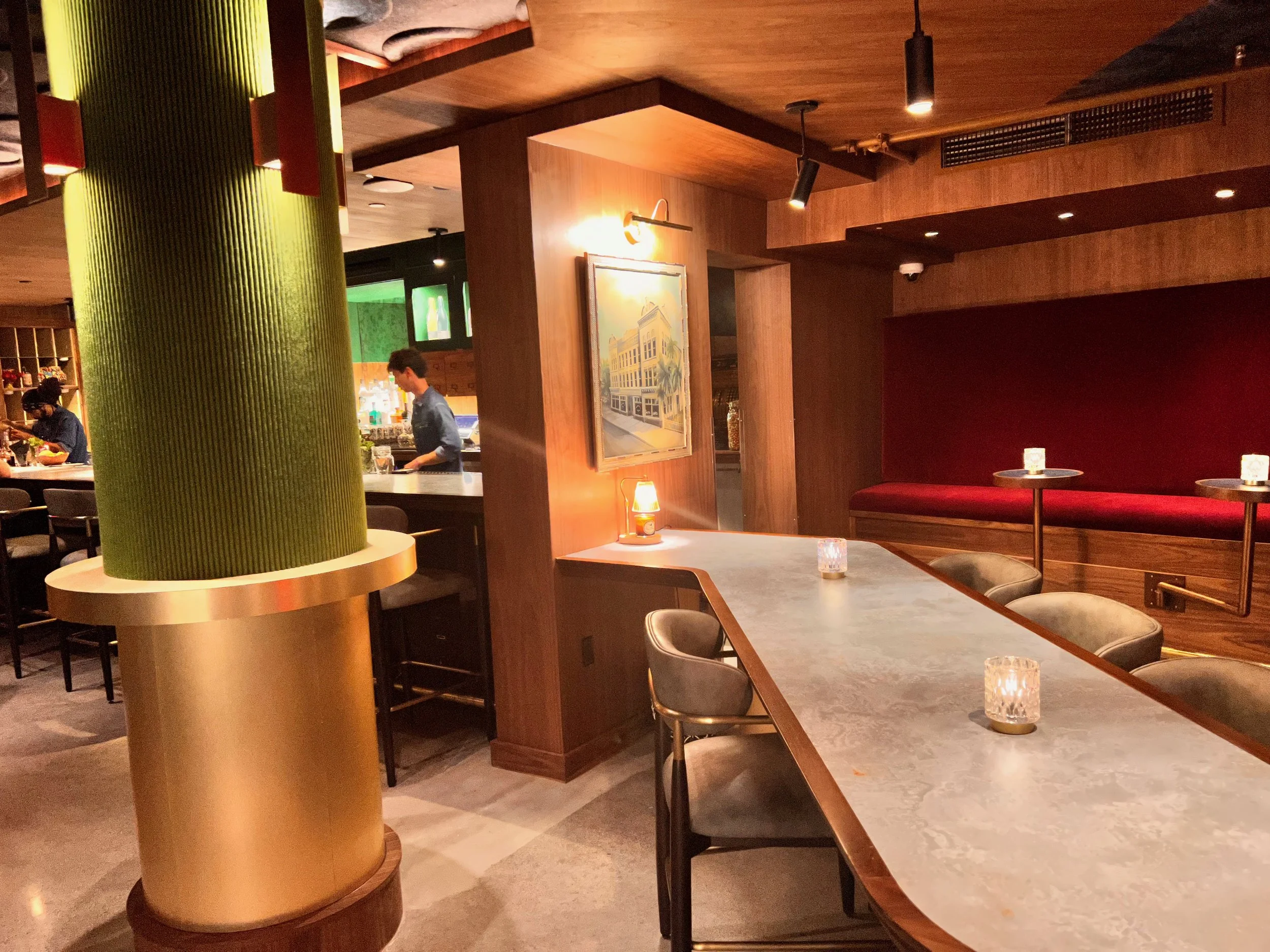 Interior of a stylish restaurant or bar with wood-paneled walls and ceiling, black lighting fixtures, a bar area in the background, and tables with candles and glassware, featuring seating with red or neutral-colored seating and a large painting on the wall.