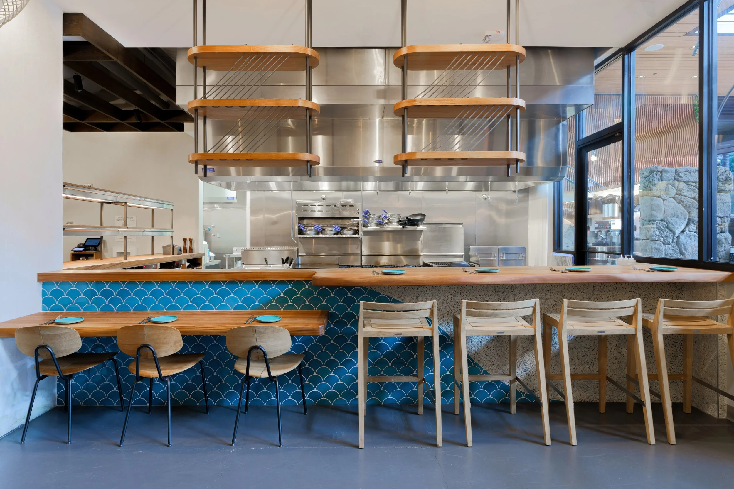 Interior view of a modern restaurant or cafe with a wooden bar counter, blue patterned tiles, and wooden chairs, stainless steel kitchen area behind the bar, large glass windows showing outdoor rocky landscape.