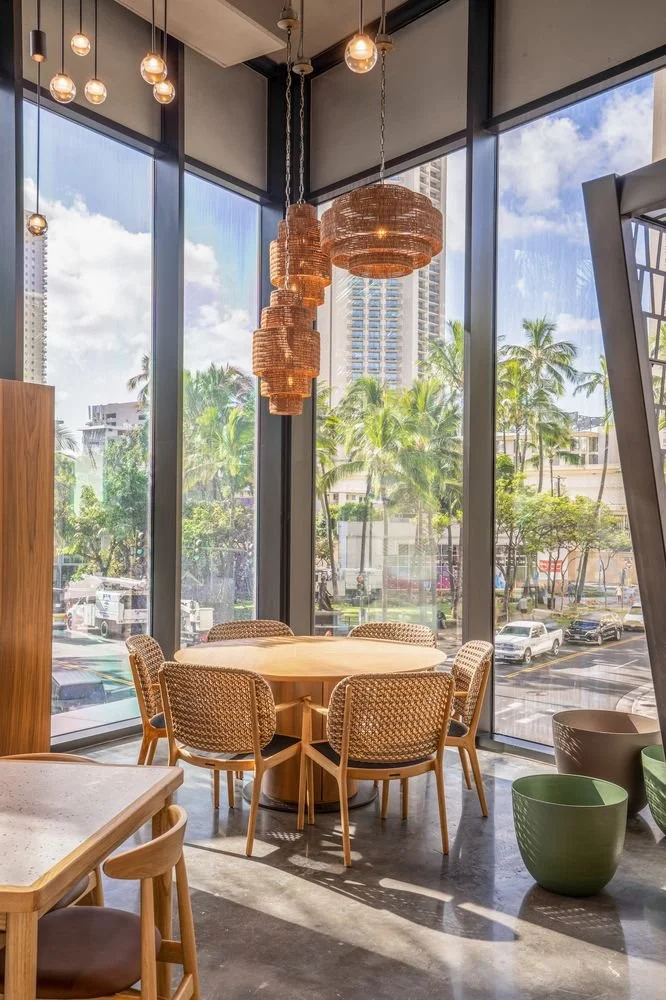 Interior of a bright dining area with a round wooden table and six woven chairs, large floor-to-ceiling windows showing palm trees and buildings outside, and decorative hanging light fixtures.