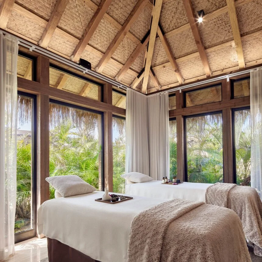 serene spa experience. couples therapy with white linens and pillows, placed under a large glass window revealing lush green plants outside, within a tropical-style bedroom with wooden framing and thatched ceiling.