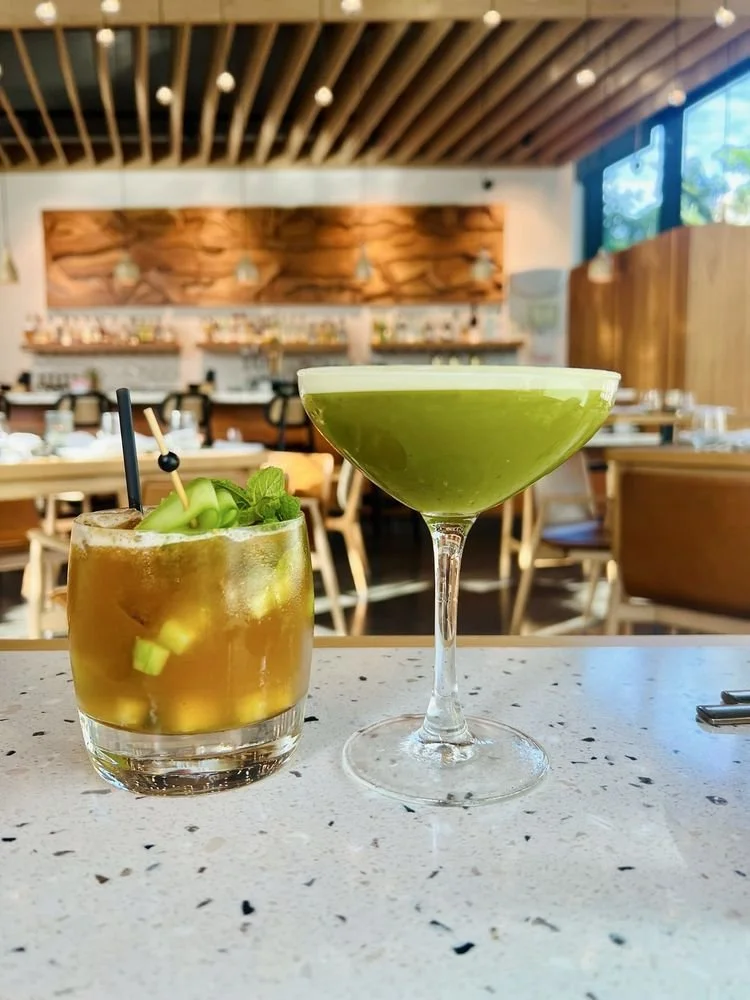 Two cocktails on a white speckled table in a restaurant: one is a brownish mixed drink with ice, cucumber, and mint garnishes in a short glass, and the other is a bright green frozen cocktail in a tall martini glass.