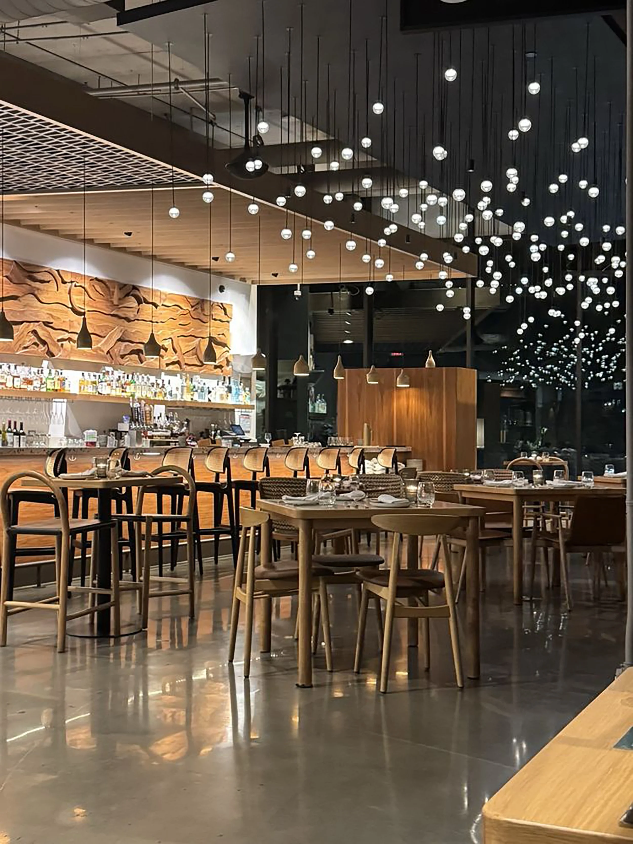 Interior of a modern restaurant or bar with wooden tables and chairs, a bar counter with bottles, stylish hanging lights, and decorative wall art.
