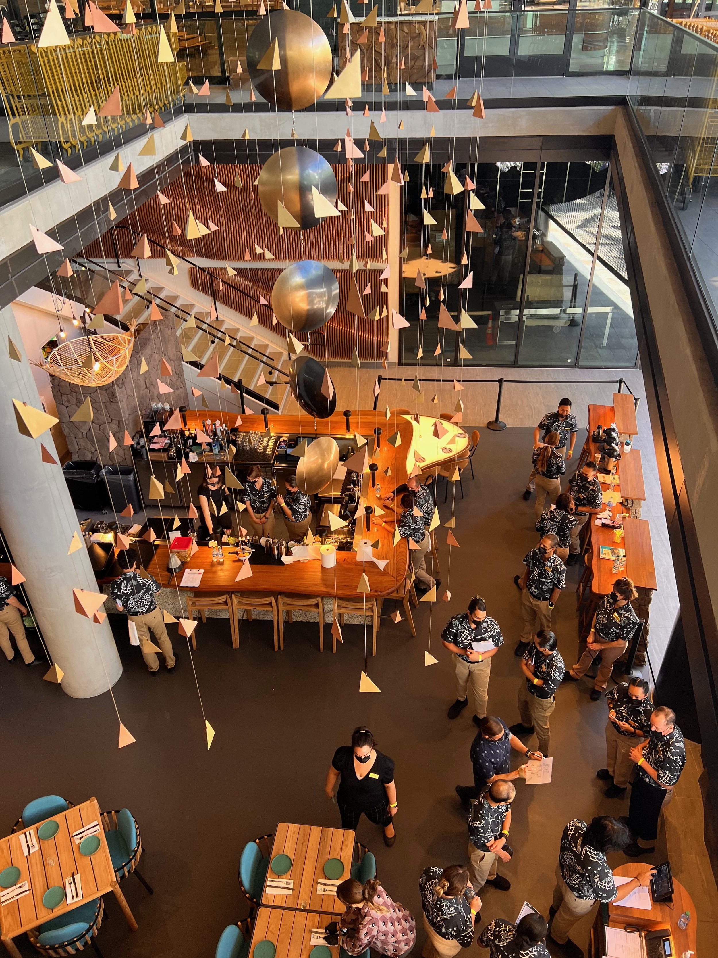 A view of a multi-level restaurant or cafe with hanging geometric decorations, a bar, tables with teal chairs, and many staff members and customers wearing black and white patterned uniforms.