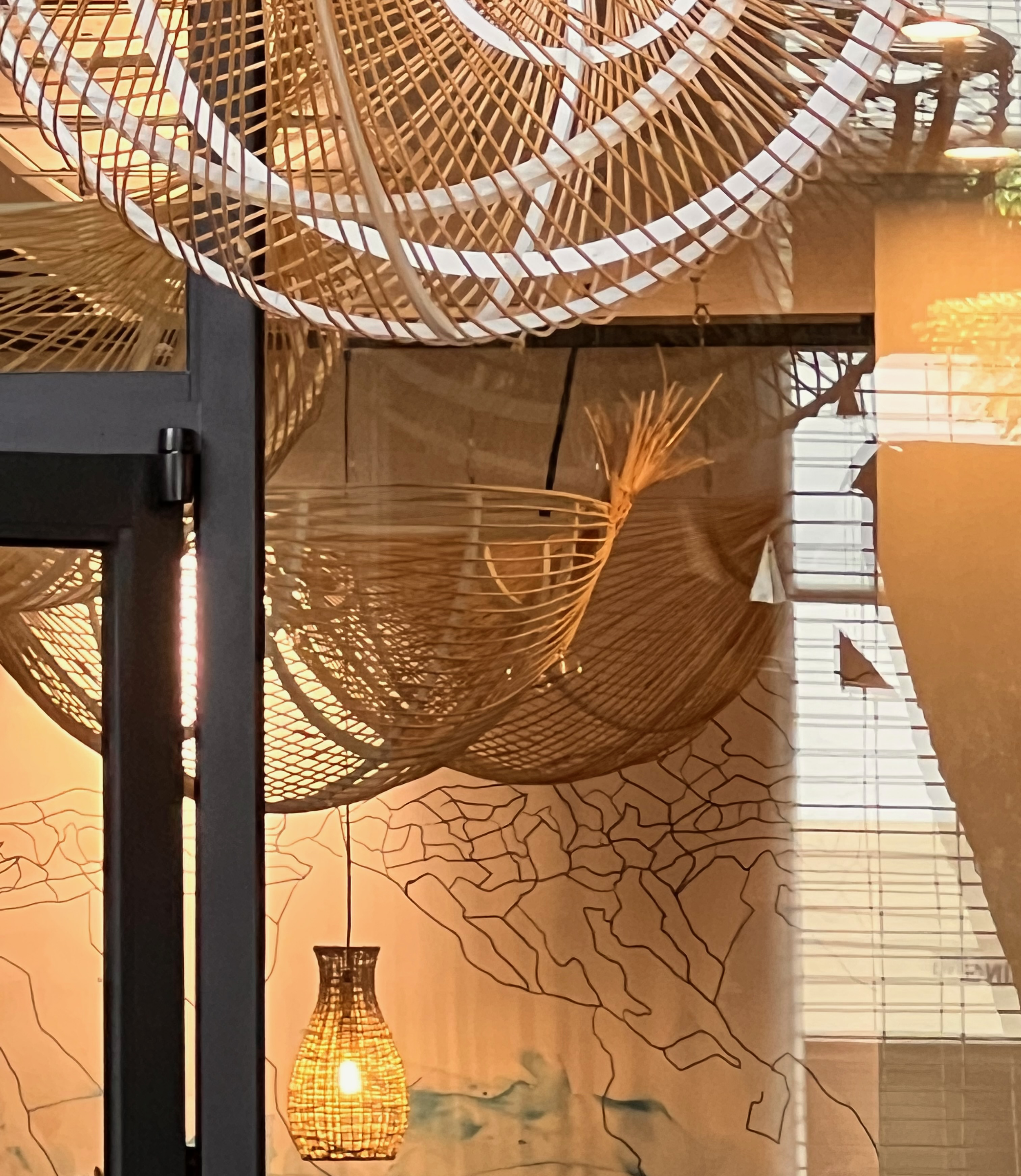 Interior of a space decorated with custom woven wicker hanging lamps and a wall with a rock wall pattern. There are sheer white curtains in front of windows, sunlight streaming through.