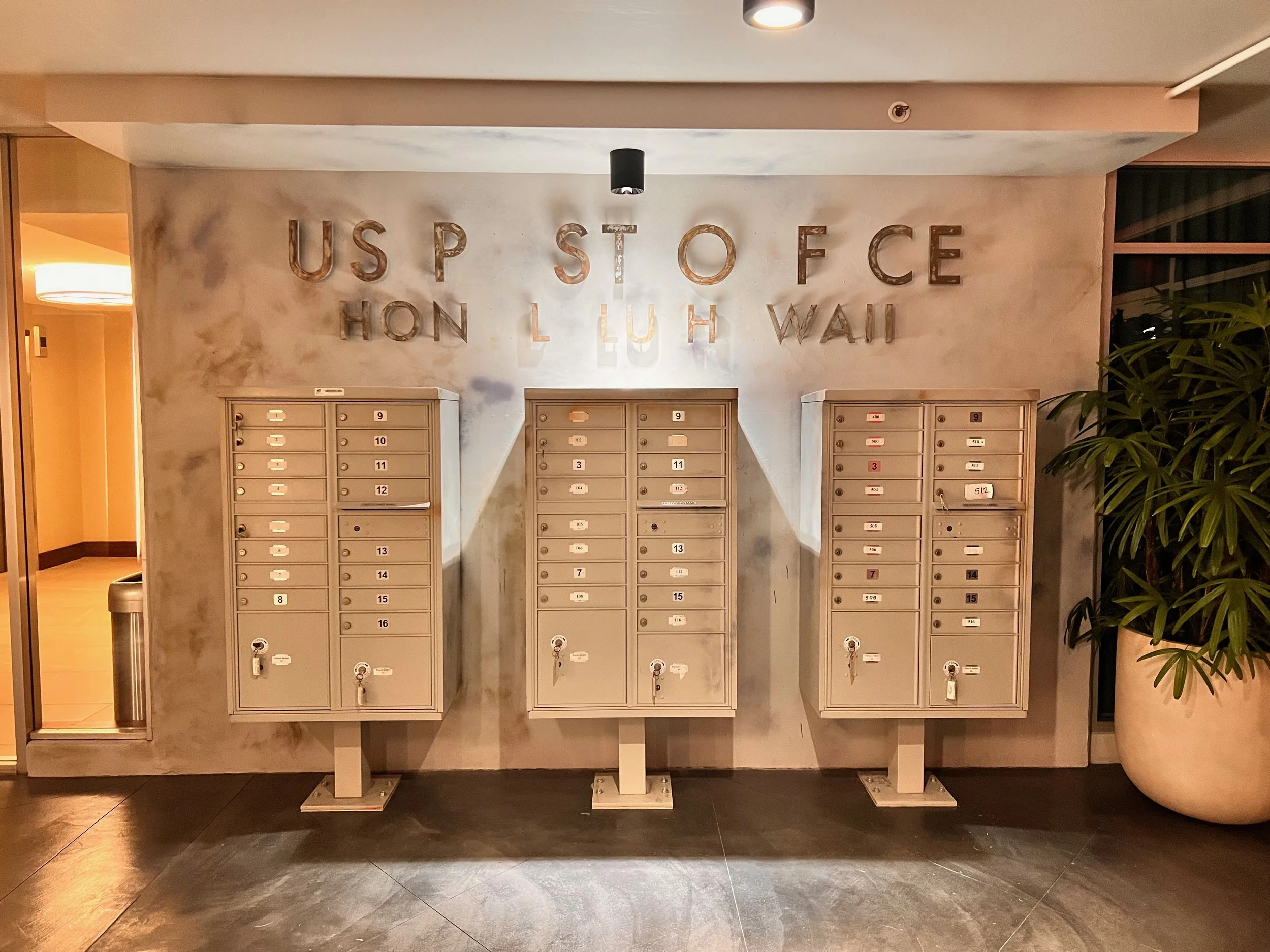 Set of beige mailboxes with numbered slots against a wall in a lobby, with a large potted plant on the right side and a wall with the words 'U.S. POST OFFICE HON L LUI H WAI' spelled out in metal letters above.