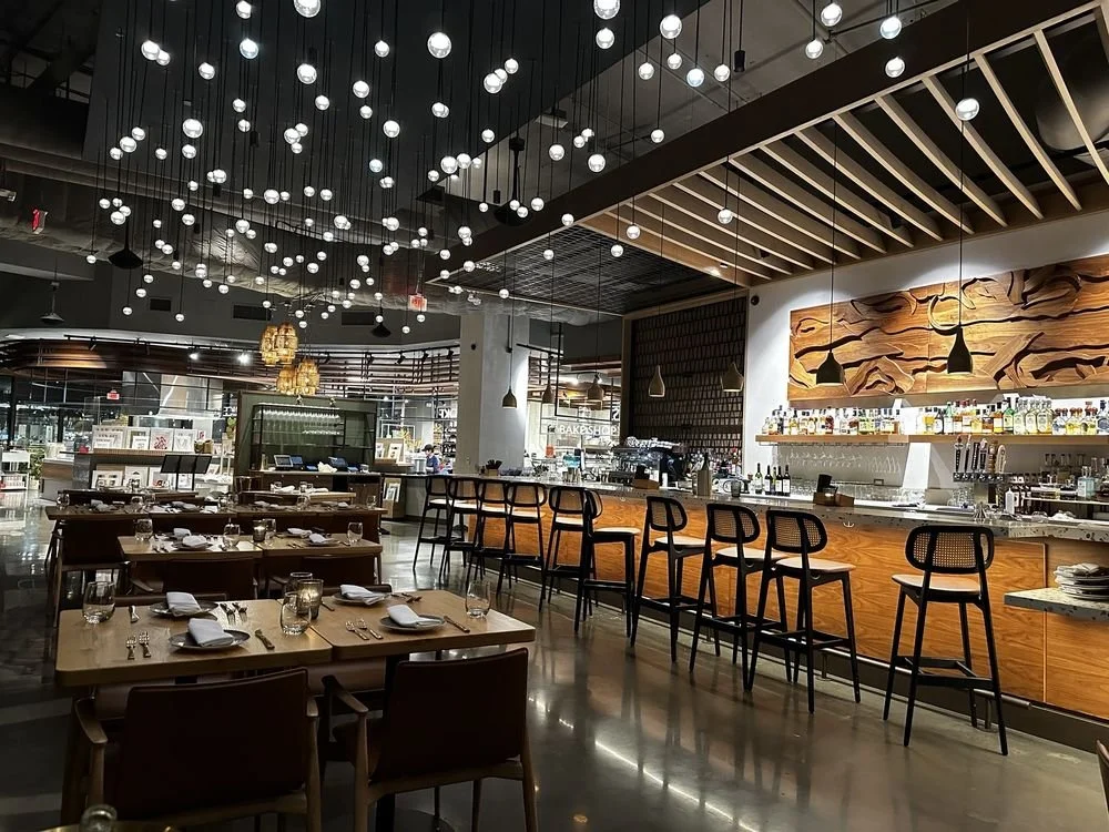 Interior of a modern restaurant or bar with wooden tables, black chairs, a bar counter with barstools, and decorative lighting fixtures on the ceiling.