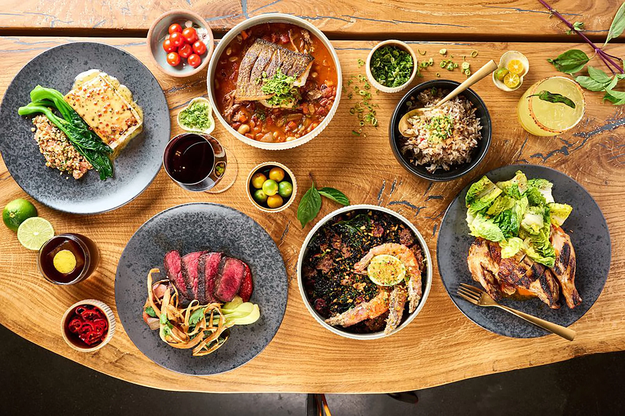 A variety of Asian dishes on a wooden table, including rice, grilled vegetables, meats, seafood, and side dishes in bowls and plates.