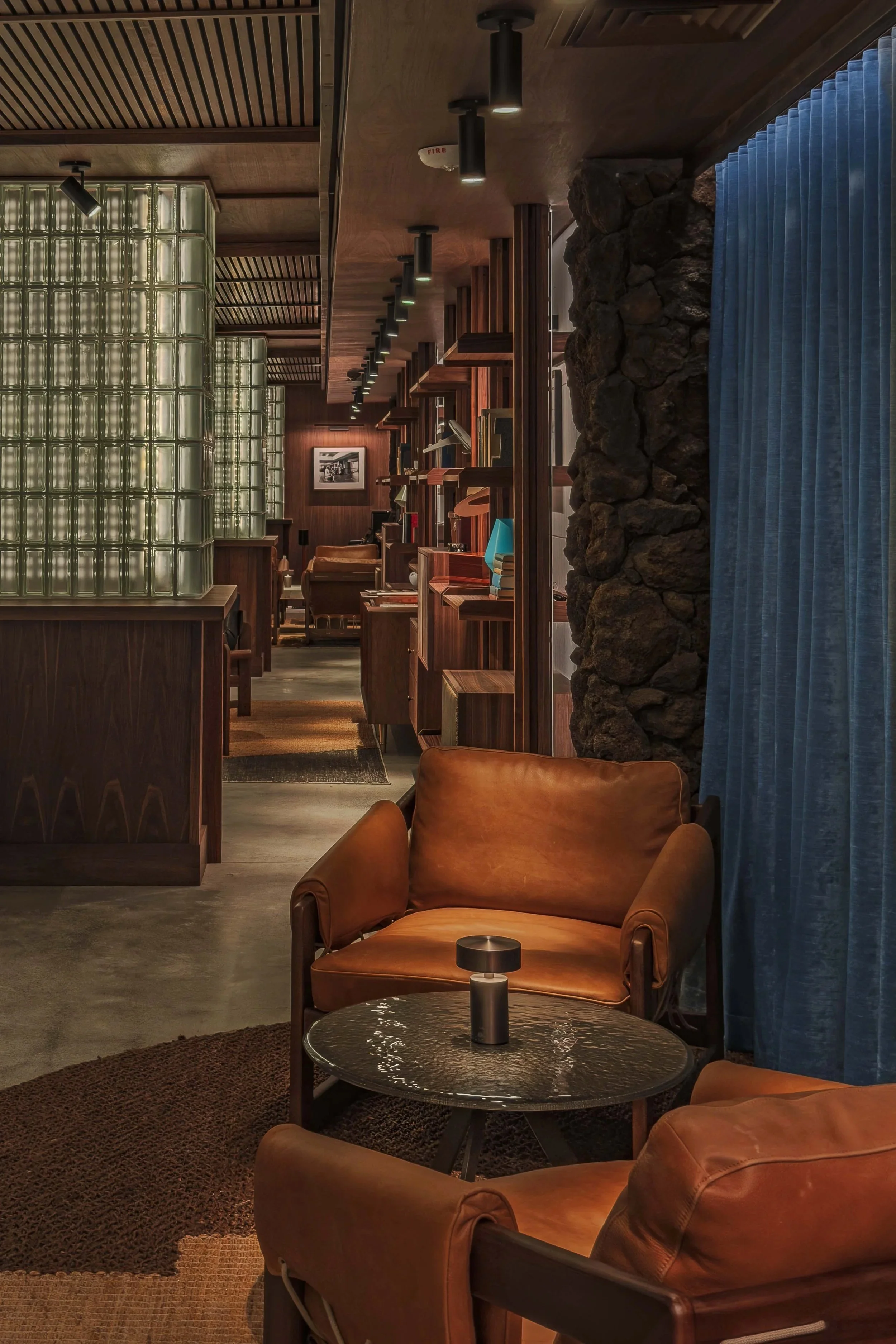 Interior of a cozy speakeasy lounge with leather armchairs, a glass block wall, wooden shelves, books, and blue curtains.