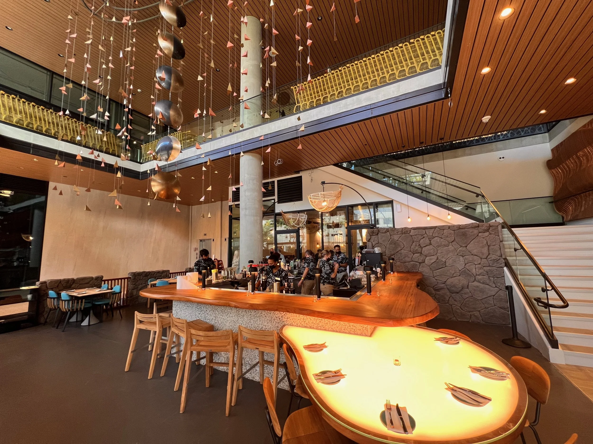 Interior of a modern restaurant with wooden ceiling and walls, hanging decorative sculptures, a large stone wall, and a bustling dining area with people dining and working.