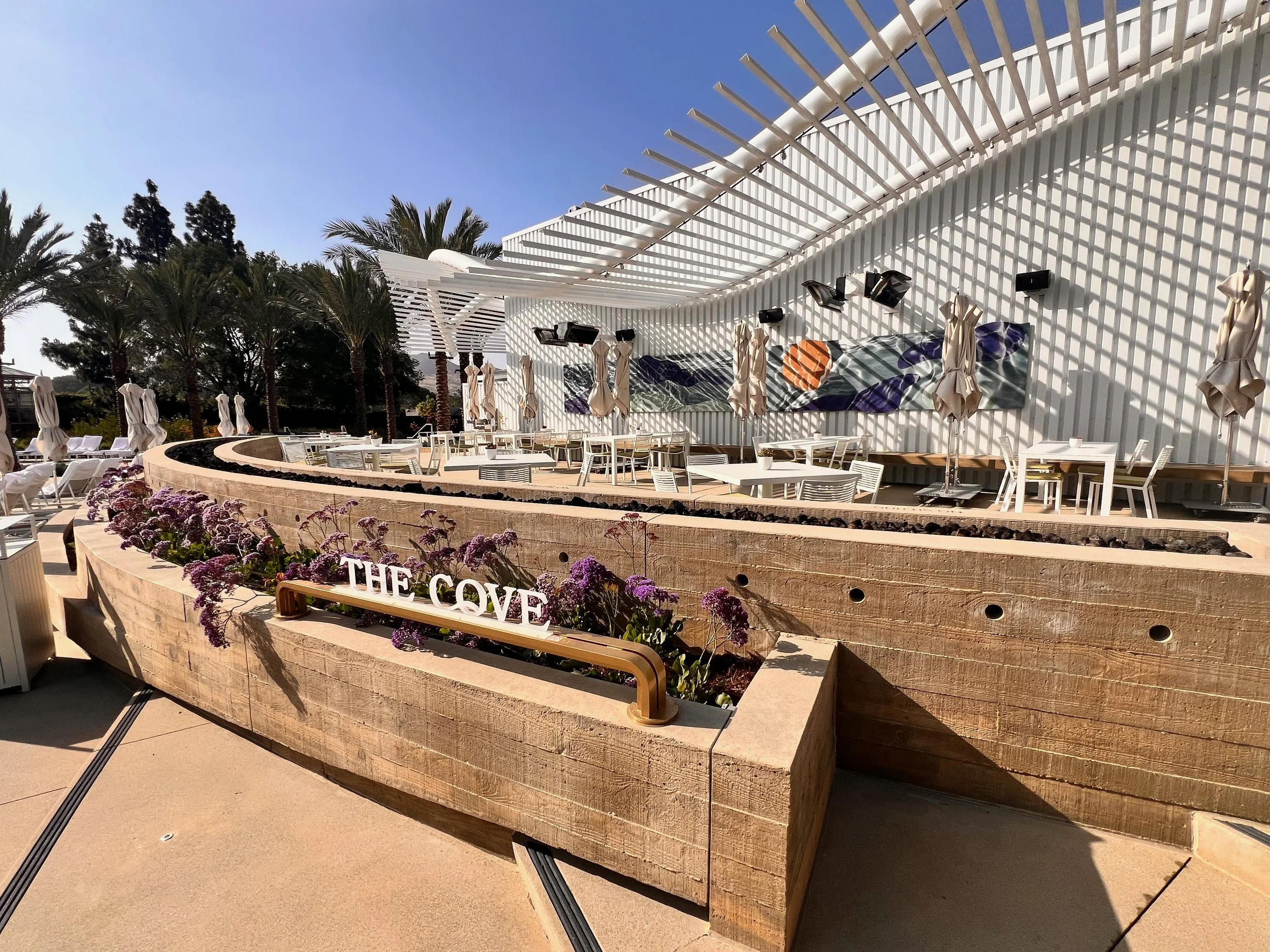 Outdoor patio at the Four Seasons Westlake with custom white tables and chairs, beige parasols, a curved wooden planter with purple flowers, a large colorful abstract banner on the wall, and tall palm with an outdoor bar with clear blue sky.