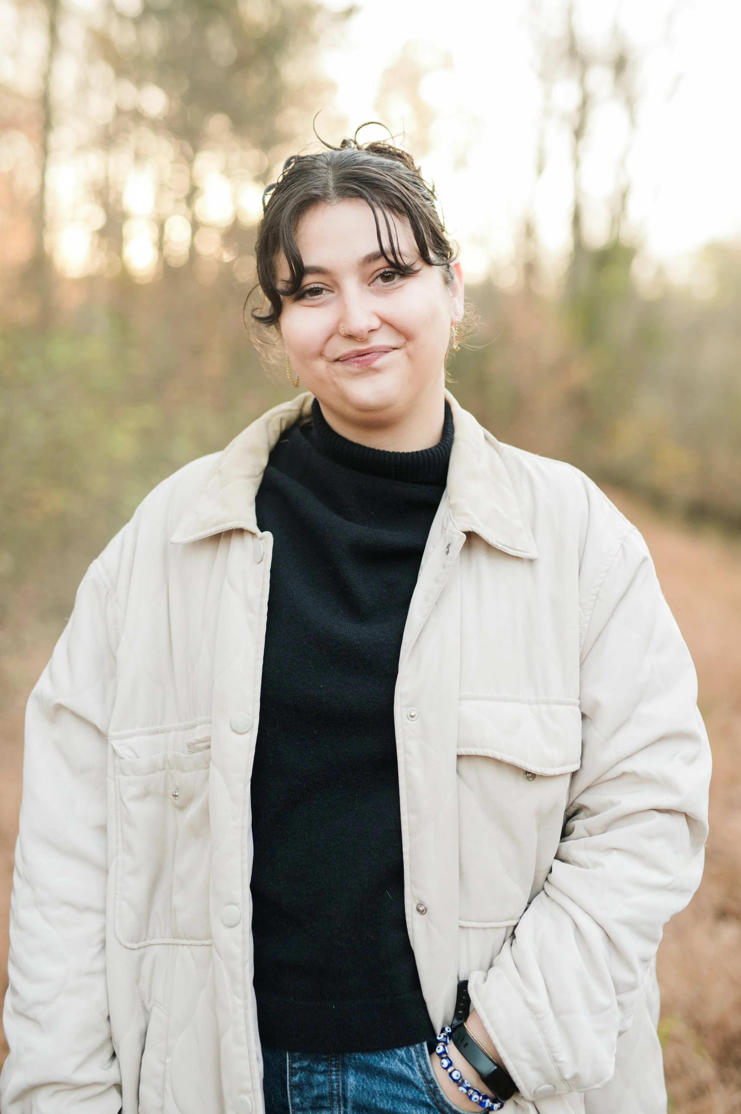 A person standing outdoors in a wooded area during fall, smiling at the camera, wearing a beige jacket over a black turtleneck, with their right hand in their pocket and a beaded bracelet.