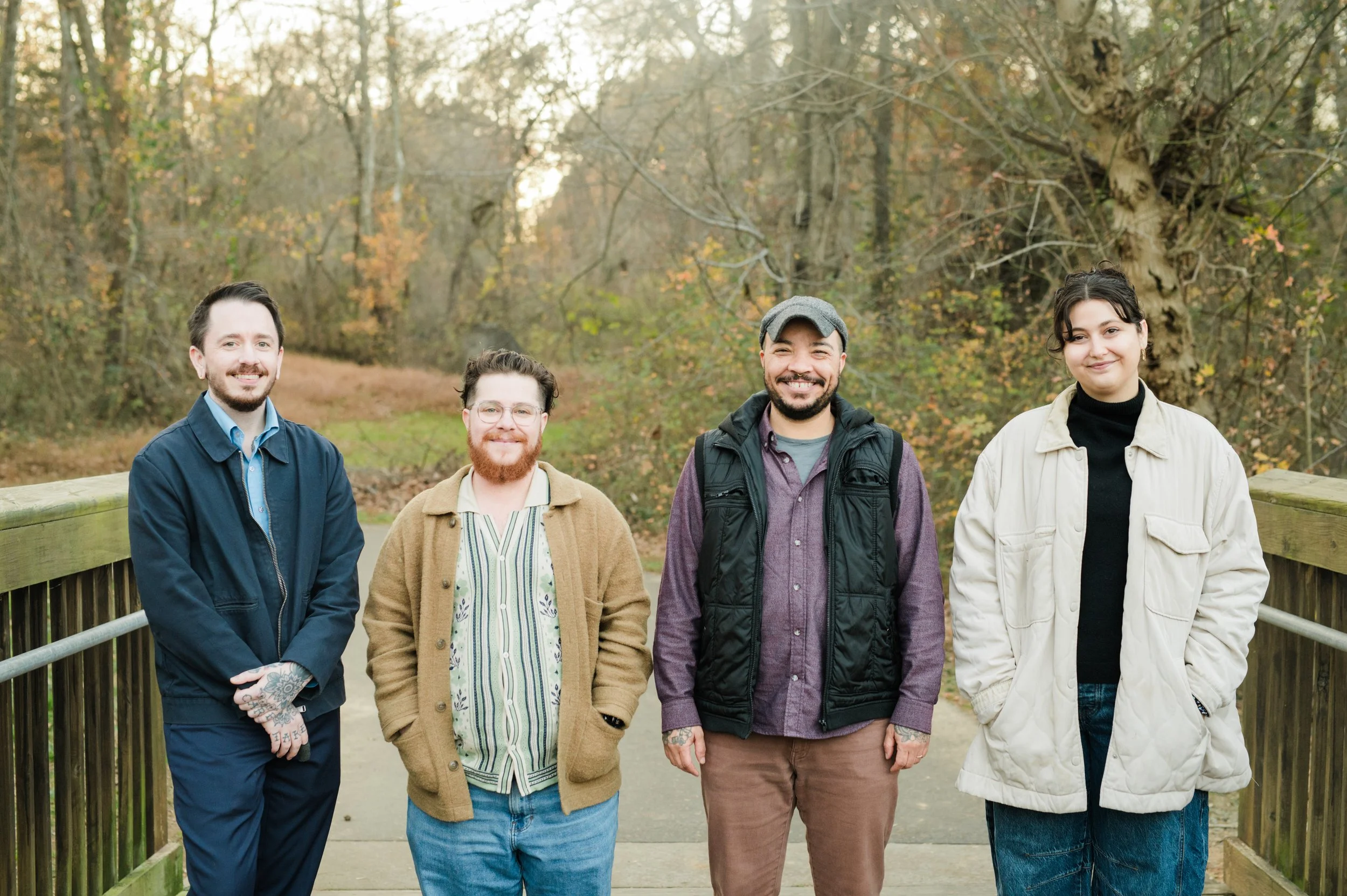 Four people standing on a wooden bridge outdoors in a forest during fall, smiling at the camera.