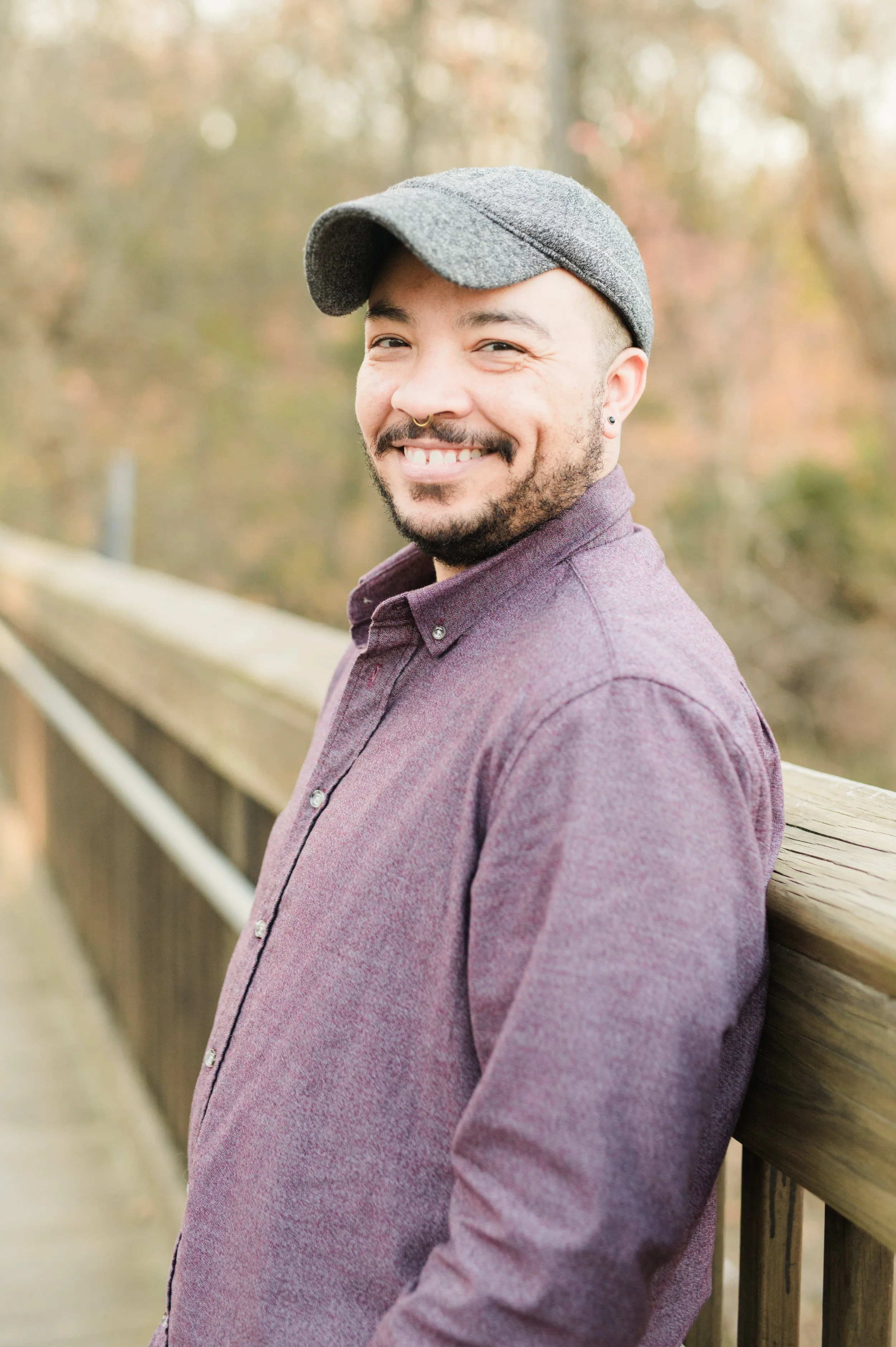 A person smiling outdoors on a wooden bridge, wearing a gray cap and a maroon shirt, with a blurred background of autumn trees.