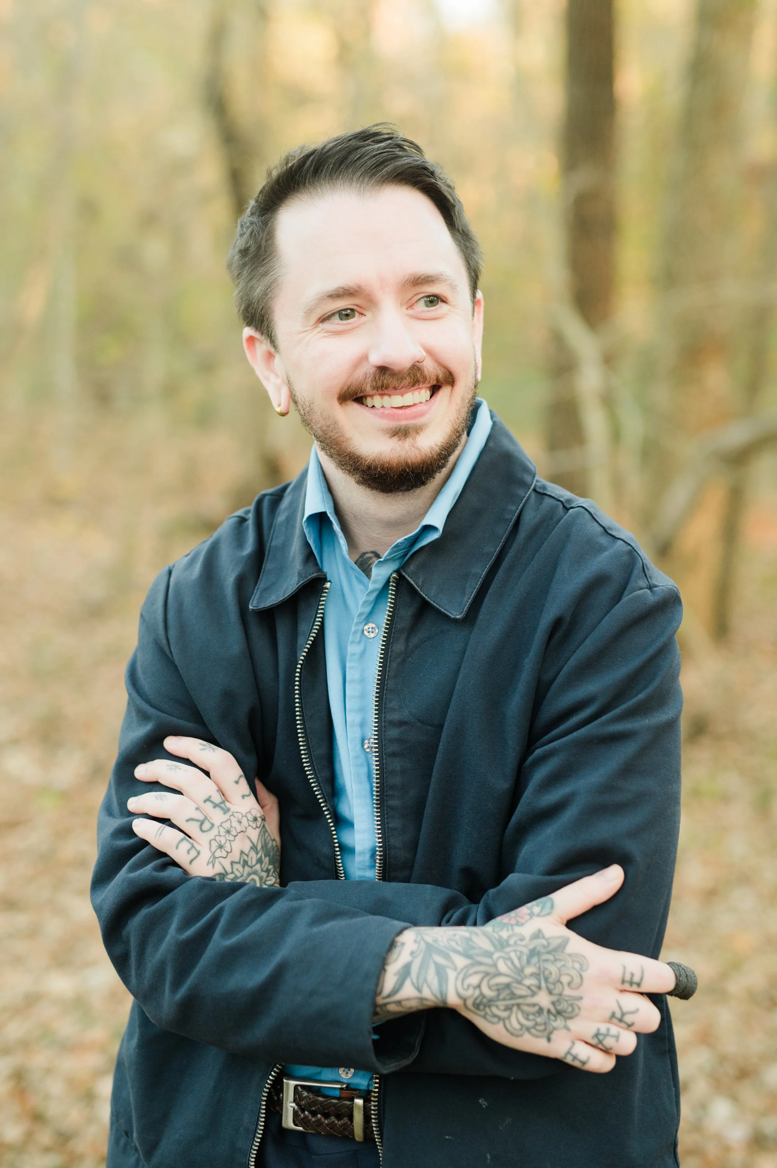 A smiling man with tattoos on his hands and arms, wearing a black jacket over a blue shirt, standing outdoors in a forest with autumn foliage.