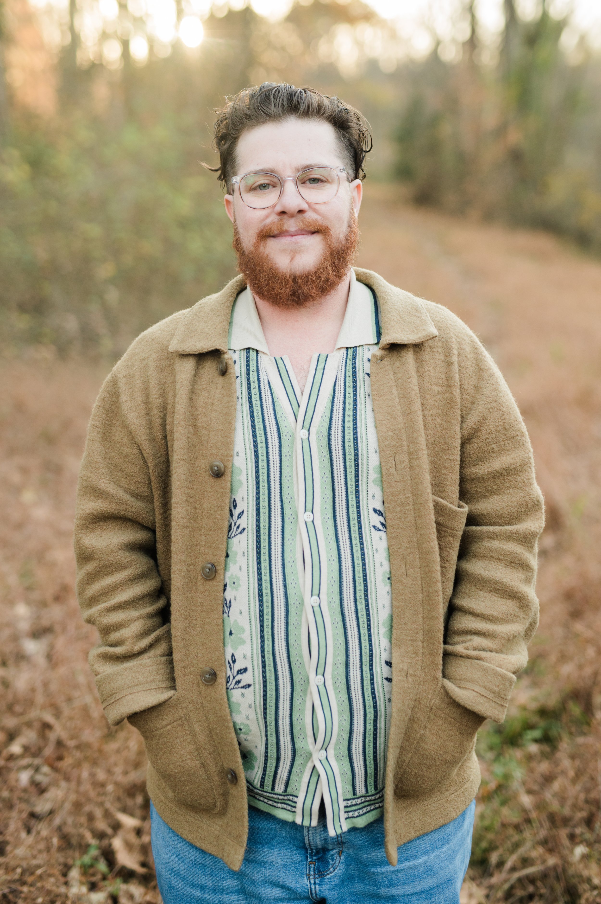 A person with glasses and a red beard standing outdoors in a forest, wearing a striped shirt, brown jacket, and jeans, with sunlight filtering through the trees.