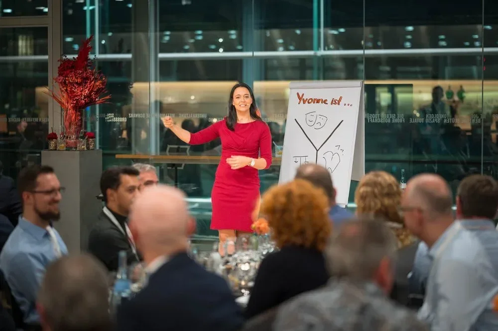 Eine Frau in einem roten Kleid hält eine Präsentation vor einem Publikum in einem modernen Veranstaltungsraum mit Glaswänden.