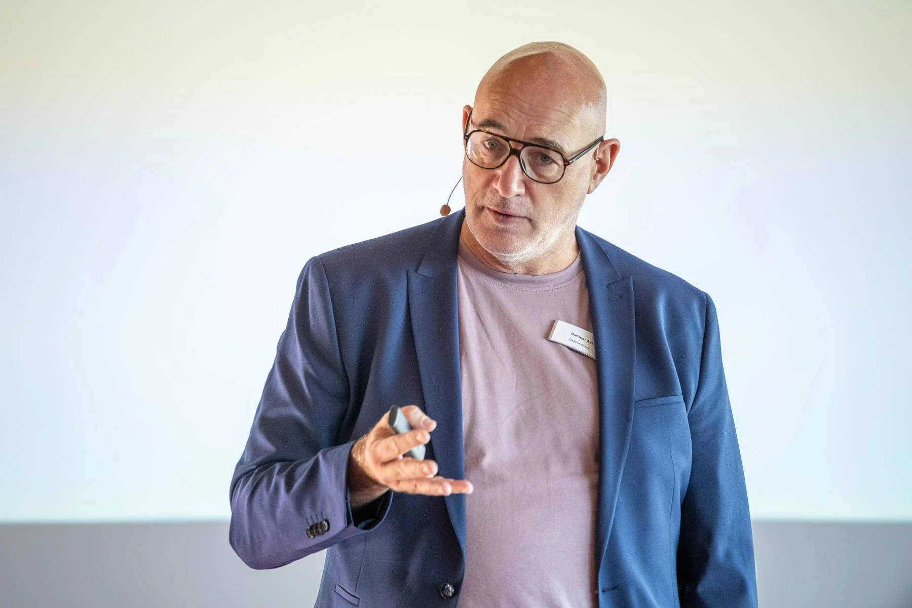 Mann mit Brille, blauer Anzug und rosa T-Shirt, spricht bei einer Präsentation, hält einen Marker in der Hand, steht vor weißer Wand.