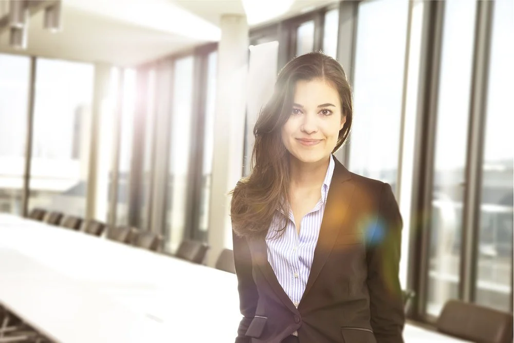 Eine junge Geschäftsfrau in einem schwarzen Blazer und Bluse vor großen Fenstern in einem modernen Büro.