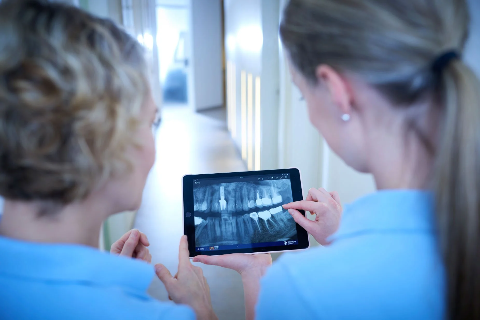 Zwei Frauen in einem Flur einer Zahnarztpraxis, die auf ein Tablet mit einer Zahn-Röntgenaufnahme schauen.